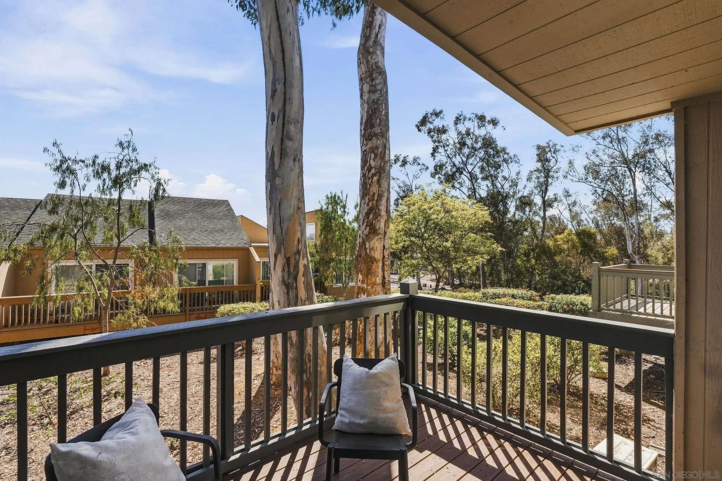 Covered wooden balcony with two cushioned chairs overlooking mature trees and a neighboring home.