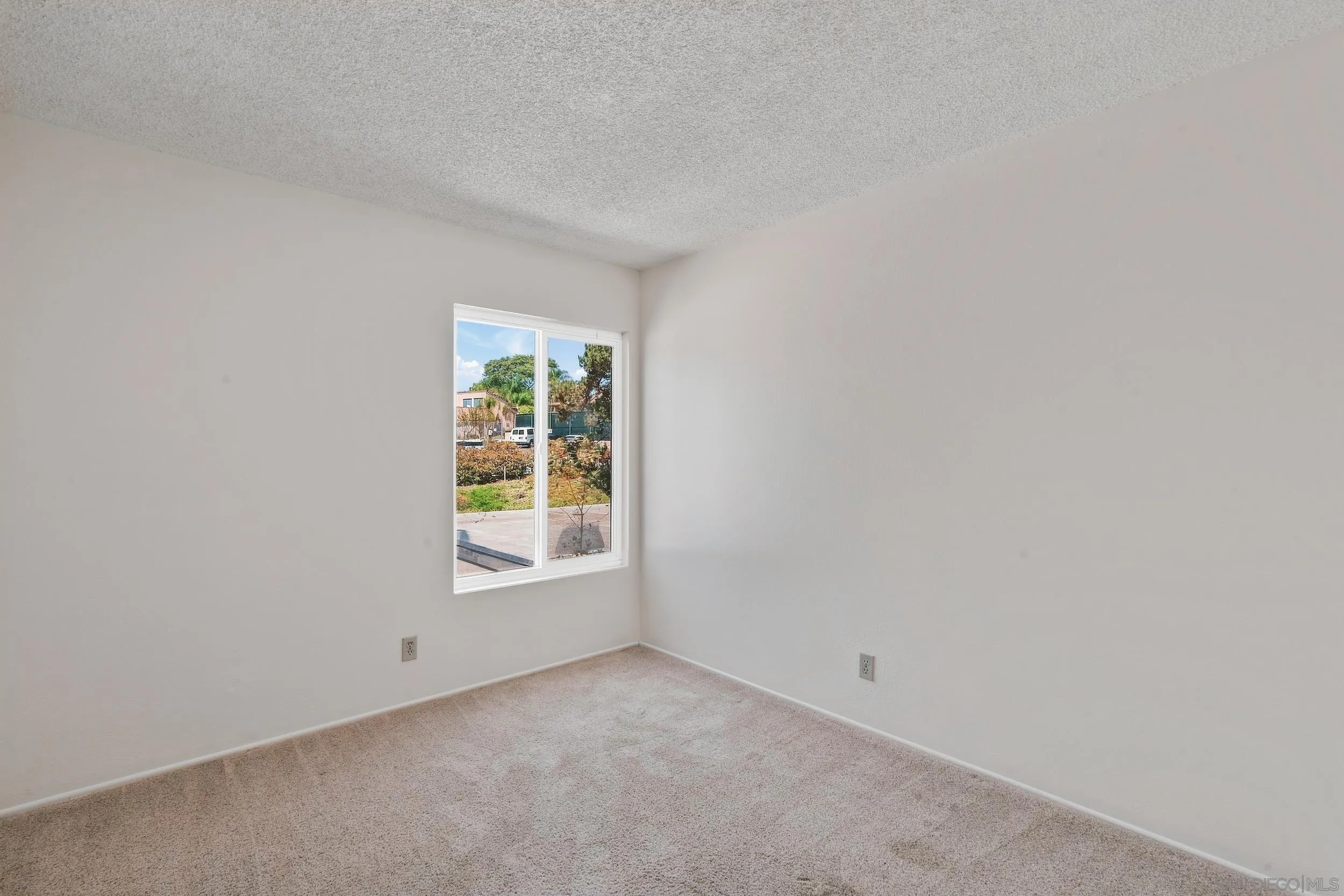 Empty carpeted room with white walls and a window overlooking greenery.