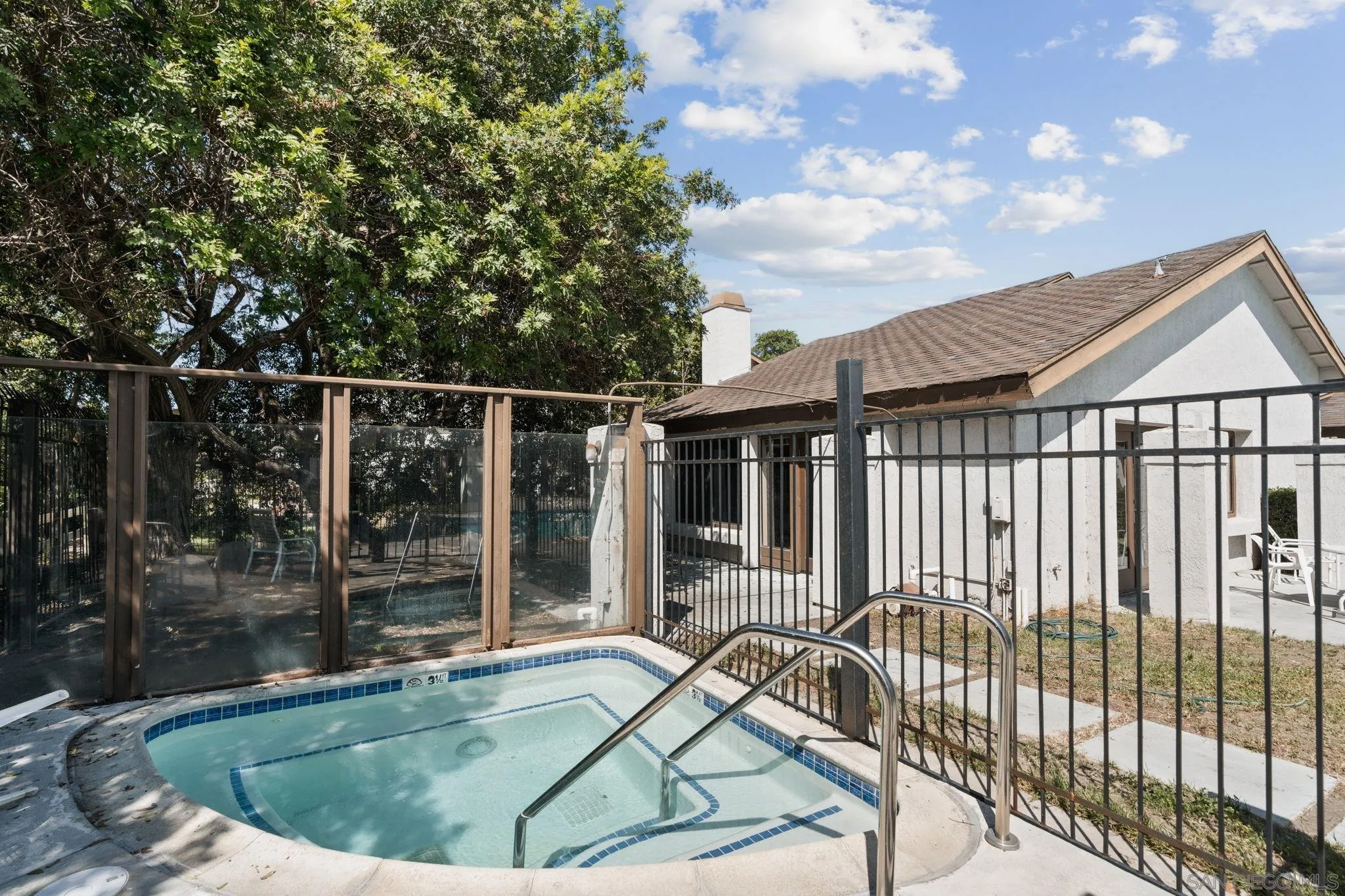 Home exterior beside a fenced tiled spa, with a concrete patio and mature trees.