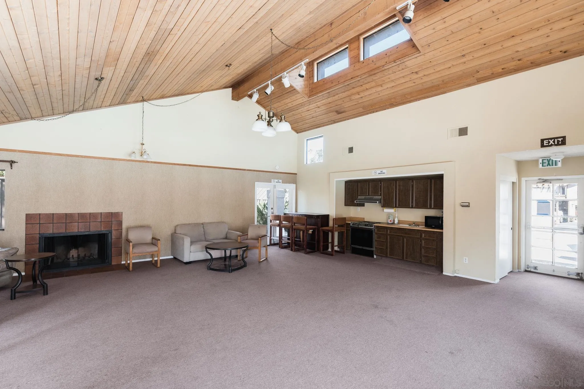 Furnished living room with a vaulted wood ceiling, brick fireplace, and open kitchen.