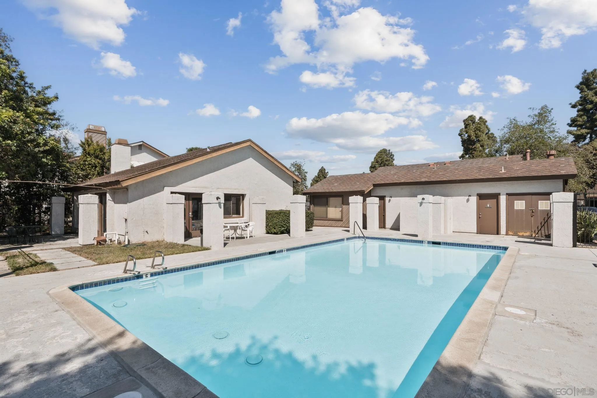 Outdoor swimming pool beside residential buildings, with a concrete deck and patio seating.