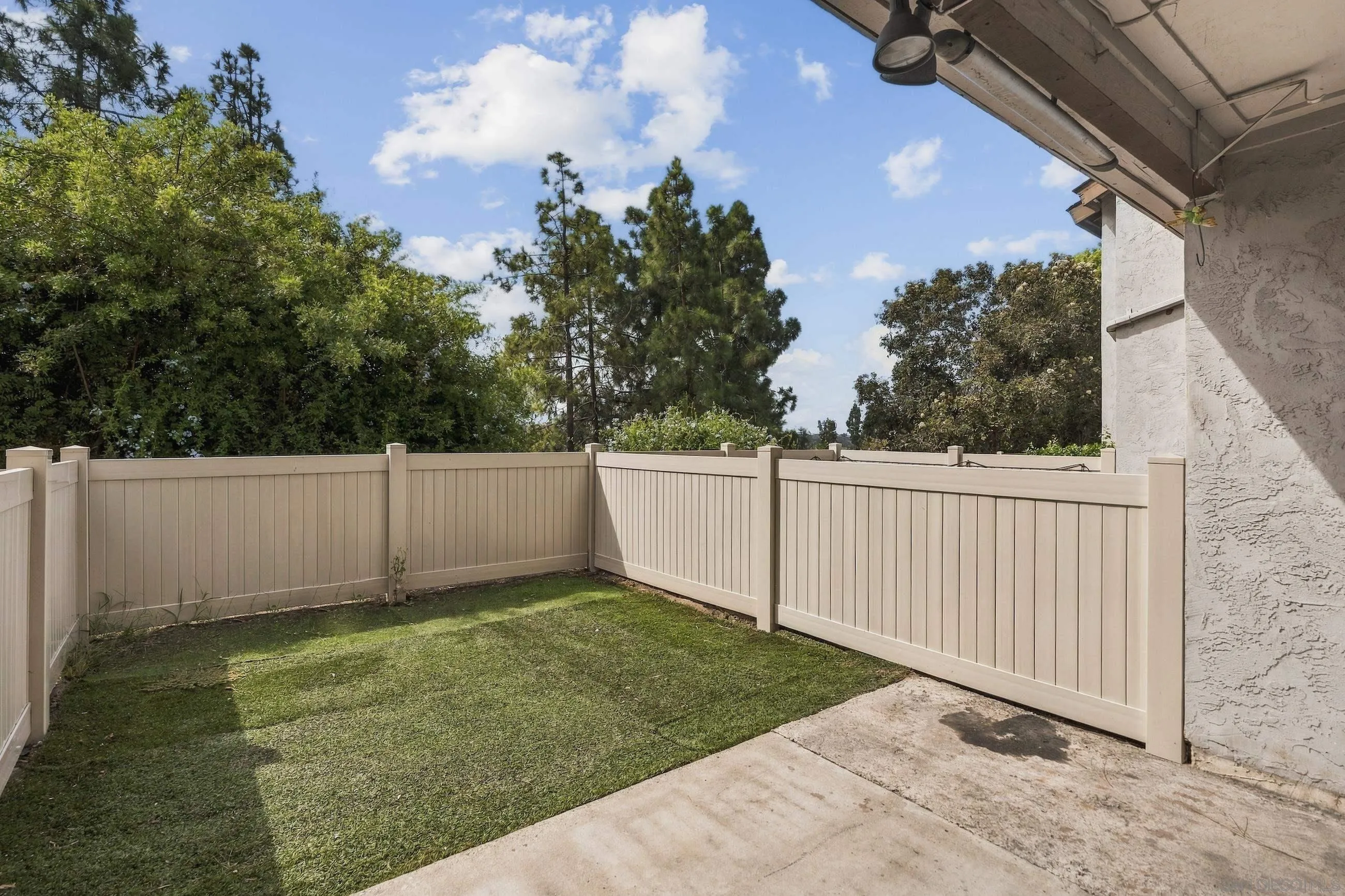 Fenced backyard with a lawn, concrete patio, and mature trees beyond the fence.