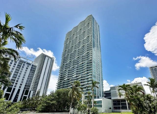 Modern glass condominium tower with curved balconies, palm trees, and neighboring high-rises.