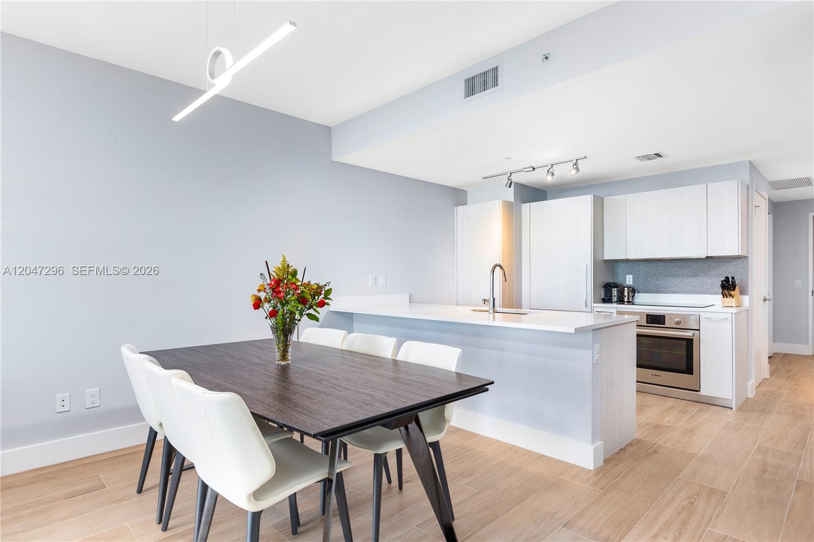 Modern condo kitchen and dining area with white cabinetry, peninsula island, and dark table with white chairs.