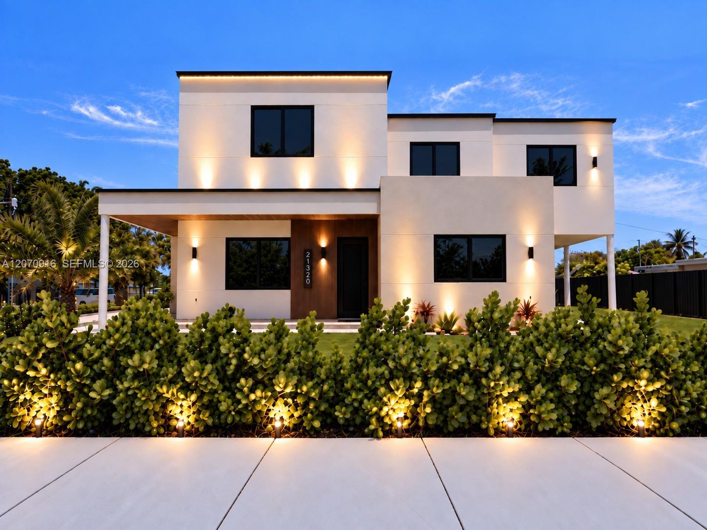 Modern two-story white home with tropical landscaping and illuminated facade at dusk.