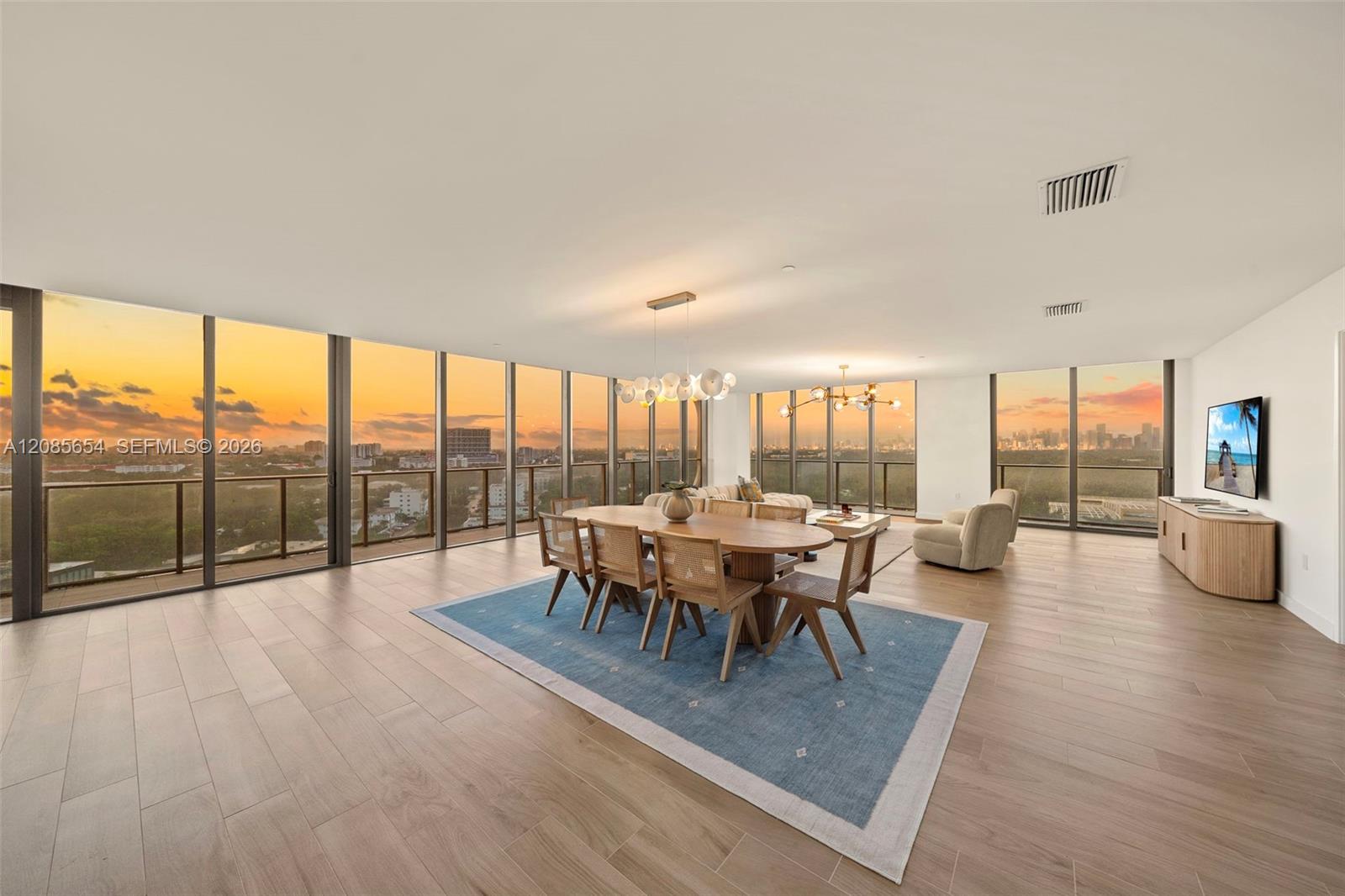 Furnished open-plan living and dining room with floor-to-ceiling windows overlooking the city at sunset.