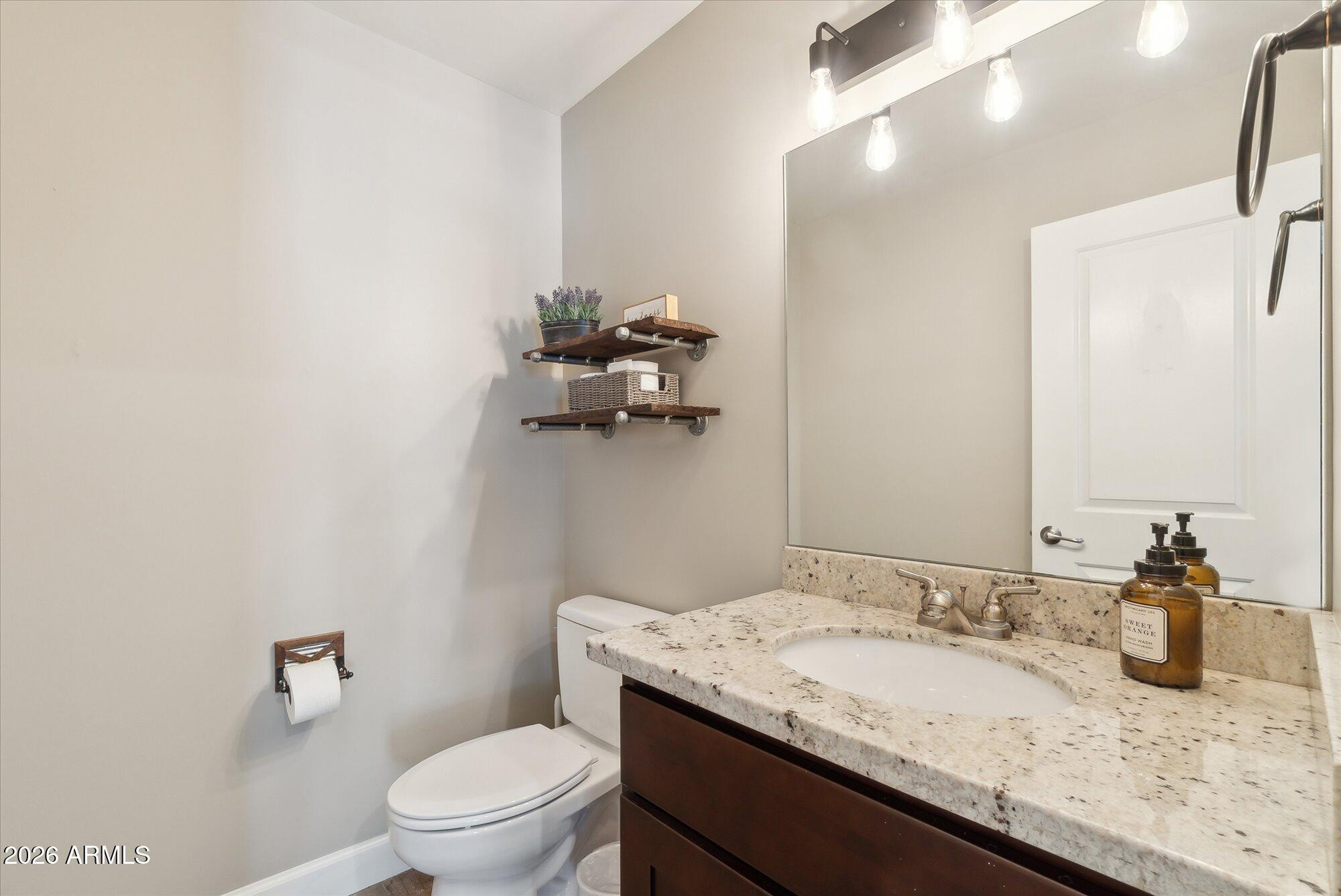 Bathroom with a granite vanity, oval sink, large mirror, and toilet beside floating shelves.
