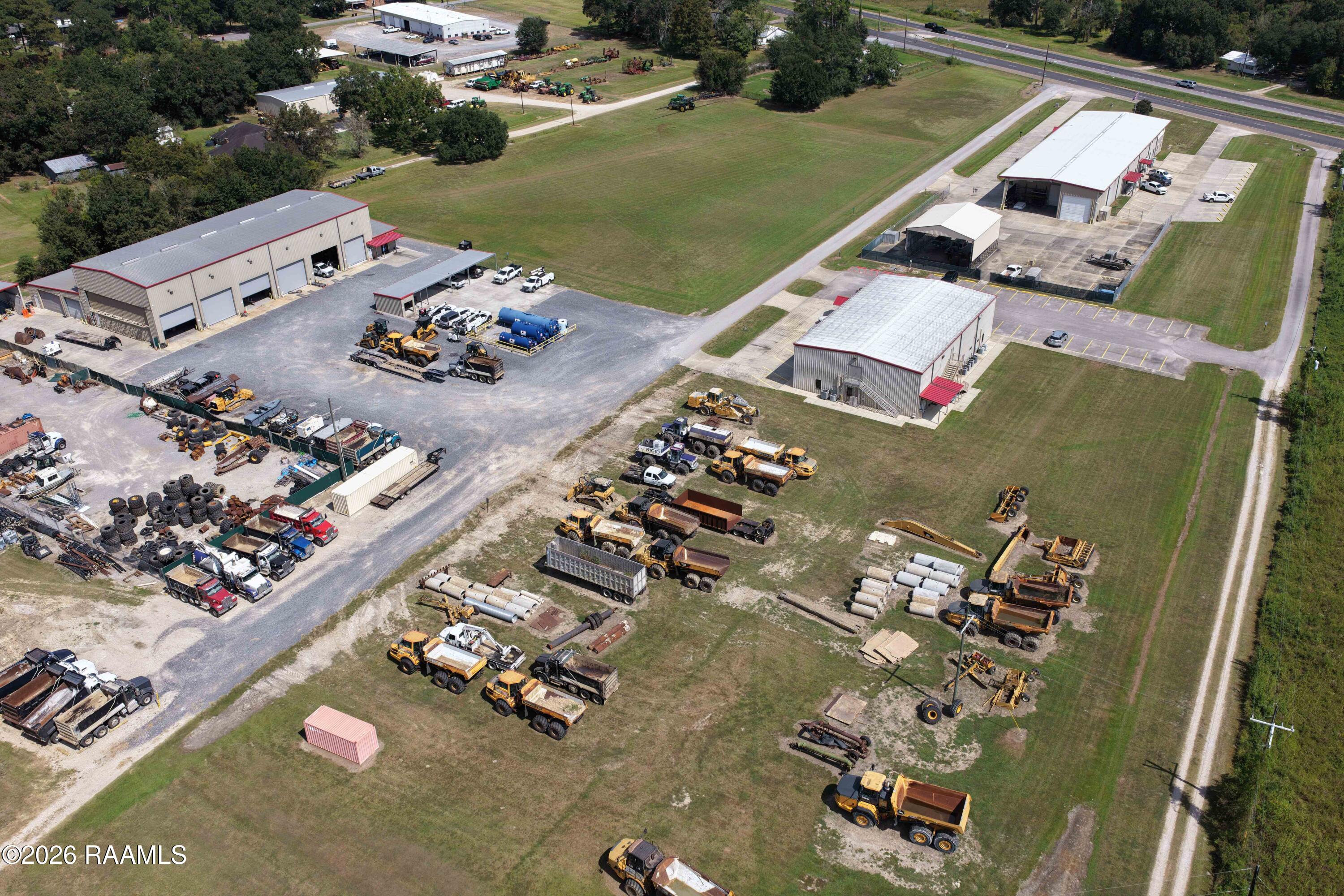 Aerial view of an equipment yard with industrial buildings, construction trucks, machinery, and open grassy land.
