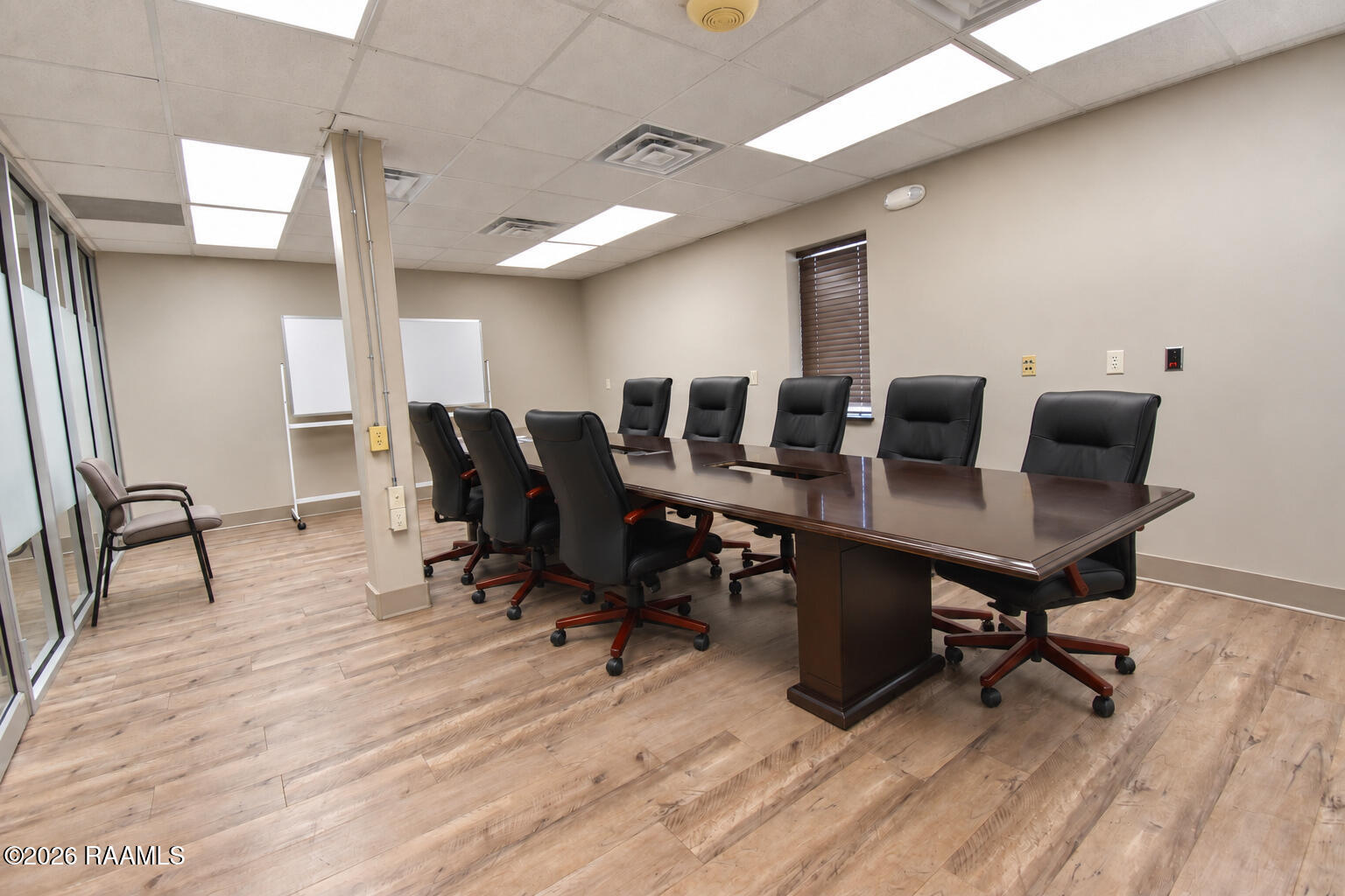 Office conference room with a long dark wood table, black chairs, glass partitions, and light wood-look flooring.