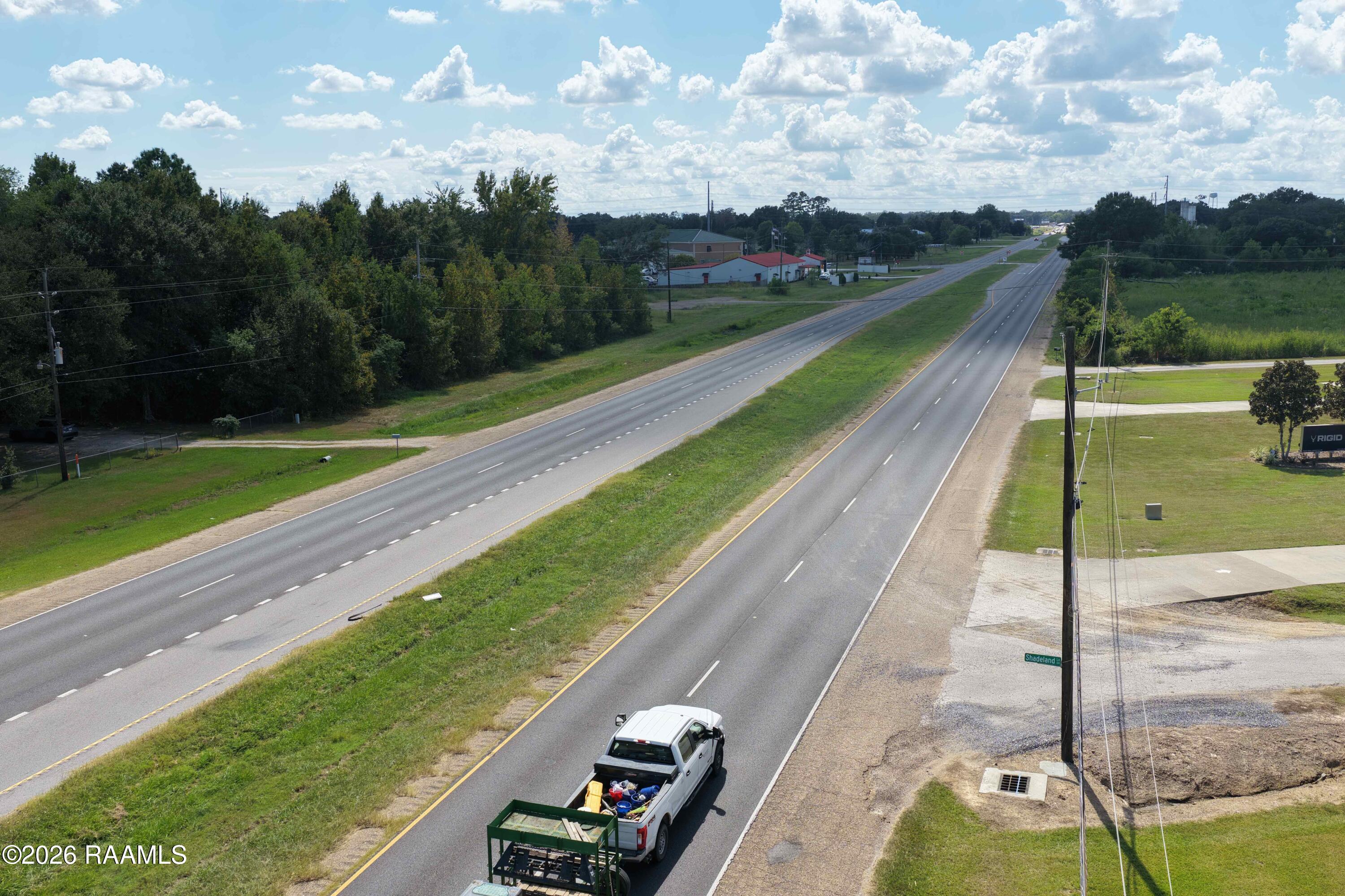 Divided highway bordered by grassy land and scattered buildings, viewed from above; a pickup towing a trailer travels nearby.