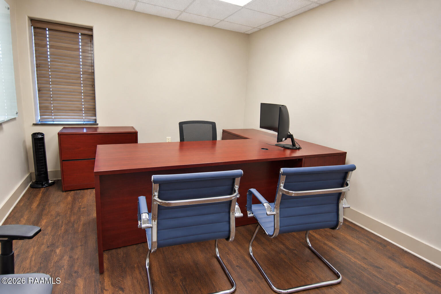 Furnished office with a large wood desk, blue visitor chairs, and a window with blinds.