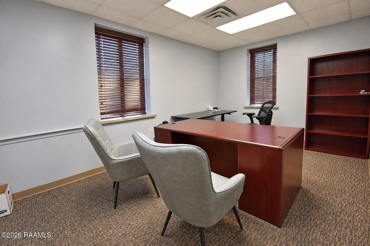 Furnished office with a reddish wood desk, gray guest chairs, and windows with dark blinds.
