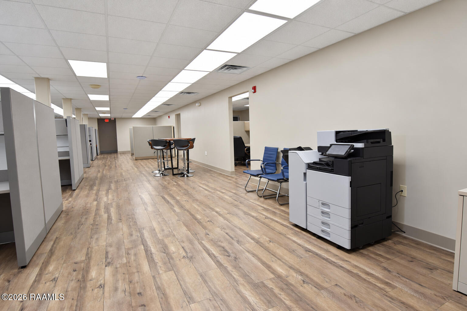 Commercial office with gray cubicles, a large copier, blue chairs, and wood-look flooring.