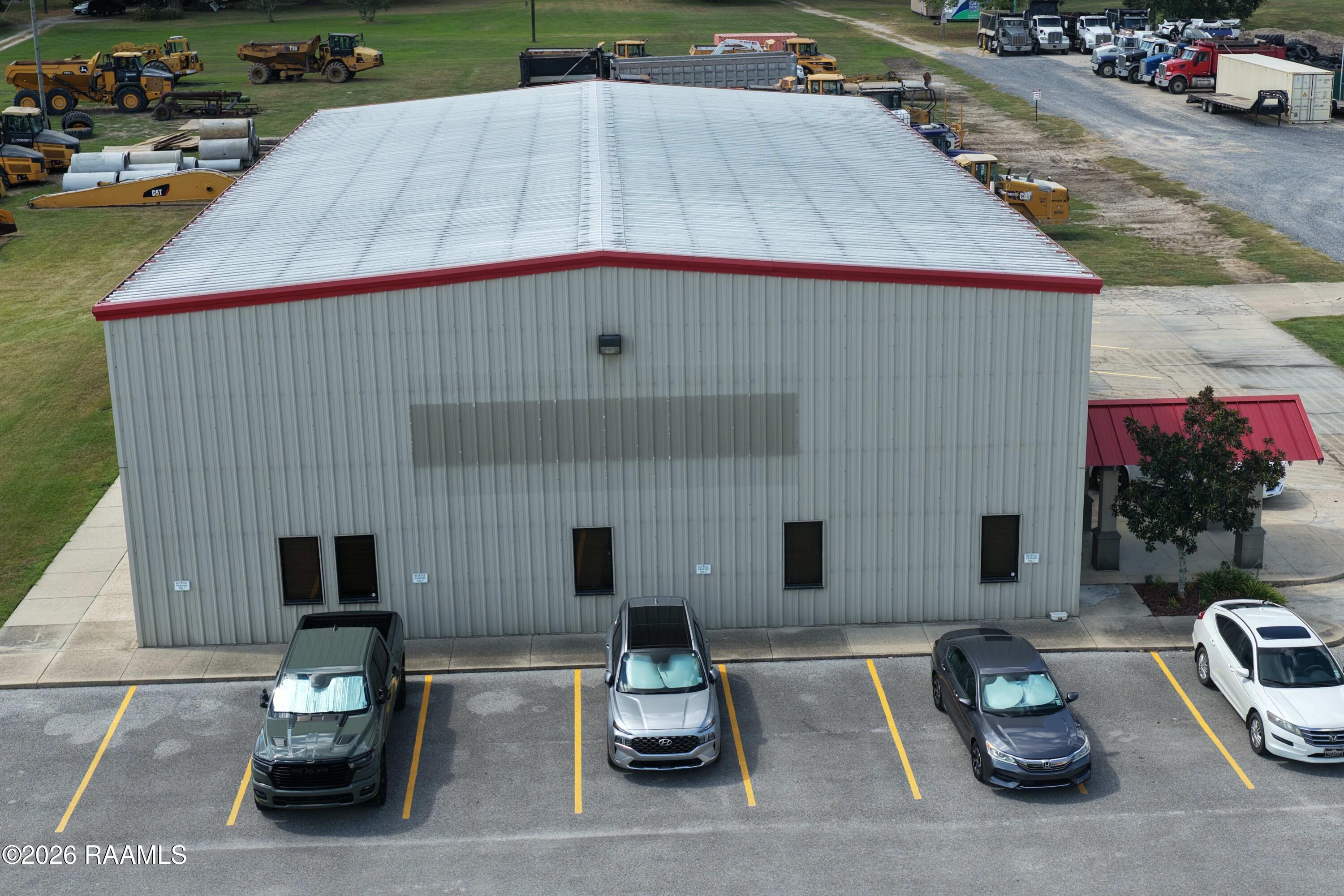 Gray metal commercial building with red roof trim, parked cars, and construction equipment in the yard.