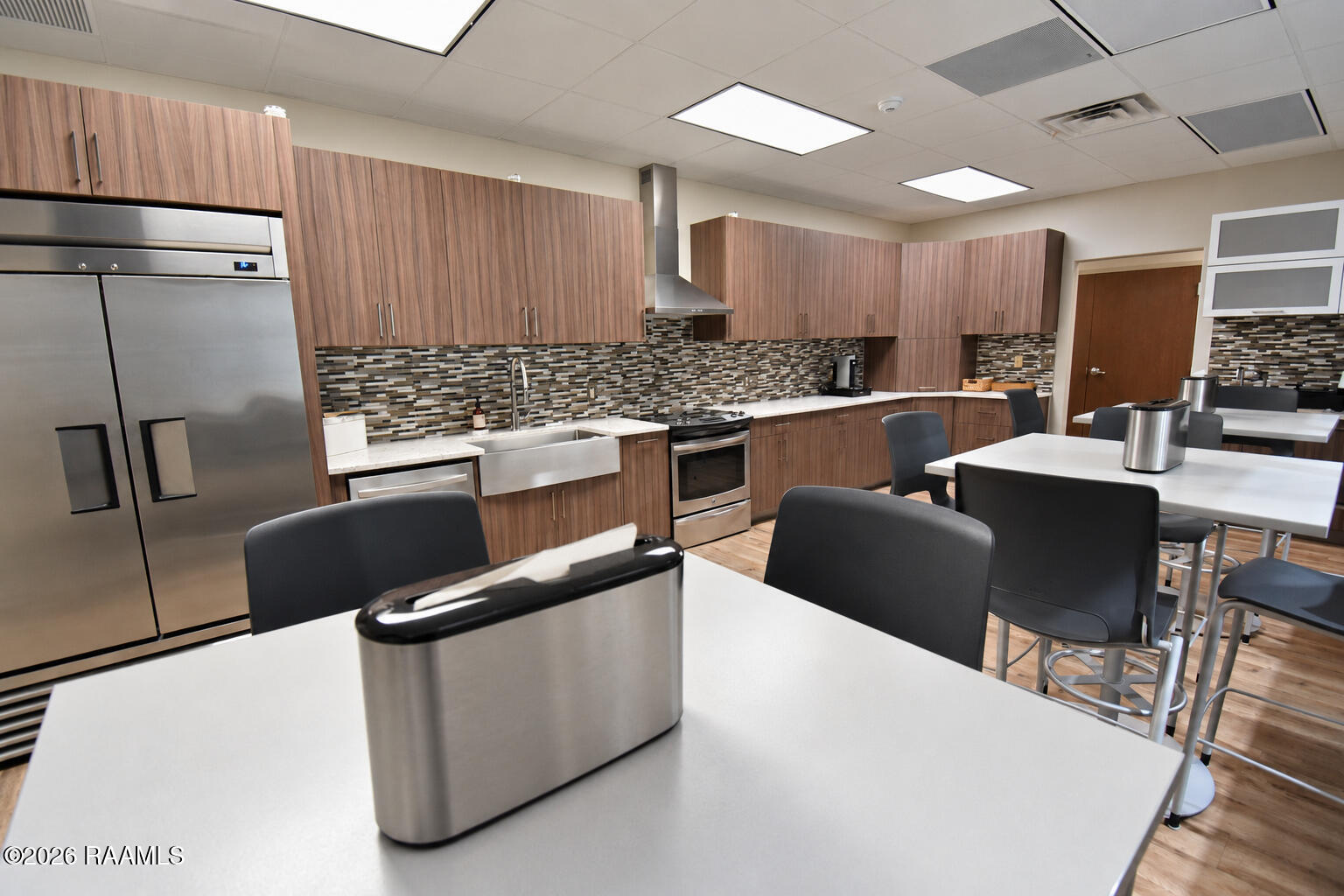 Commercial kitchen with stainless steel appliances, wood-tone cabinetry, and adjacent tables and chairs.