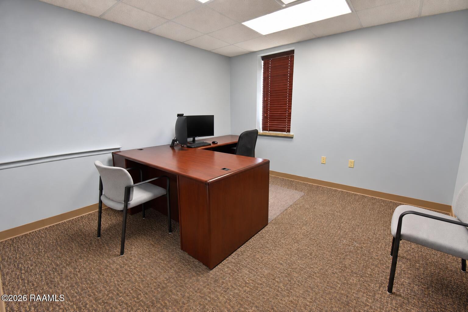 Office with a large wooden desk, computer, patterned carpet, and a narrow window with blinds.