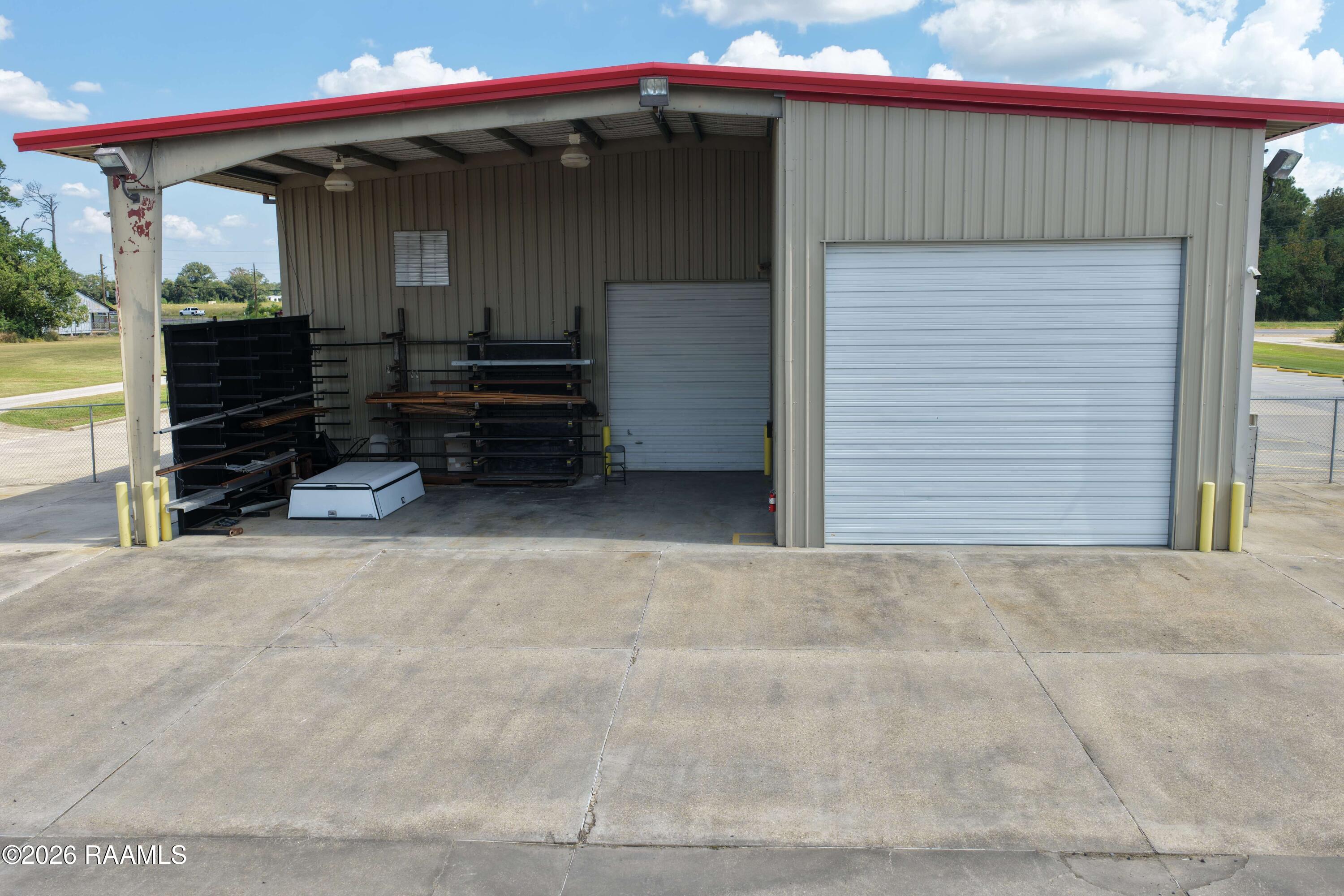 Metal workshop exterior with an open covered work bay, storage racks, and a large roll-up door.