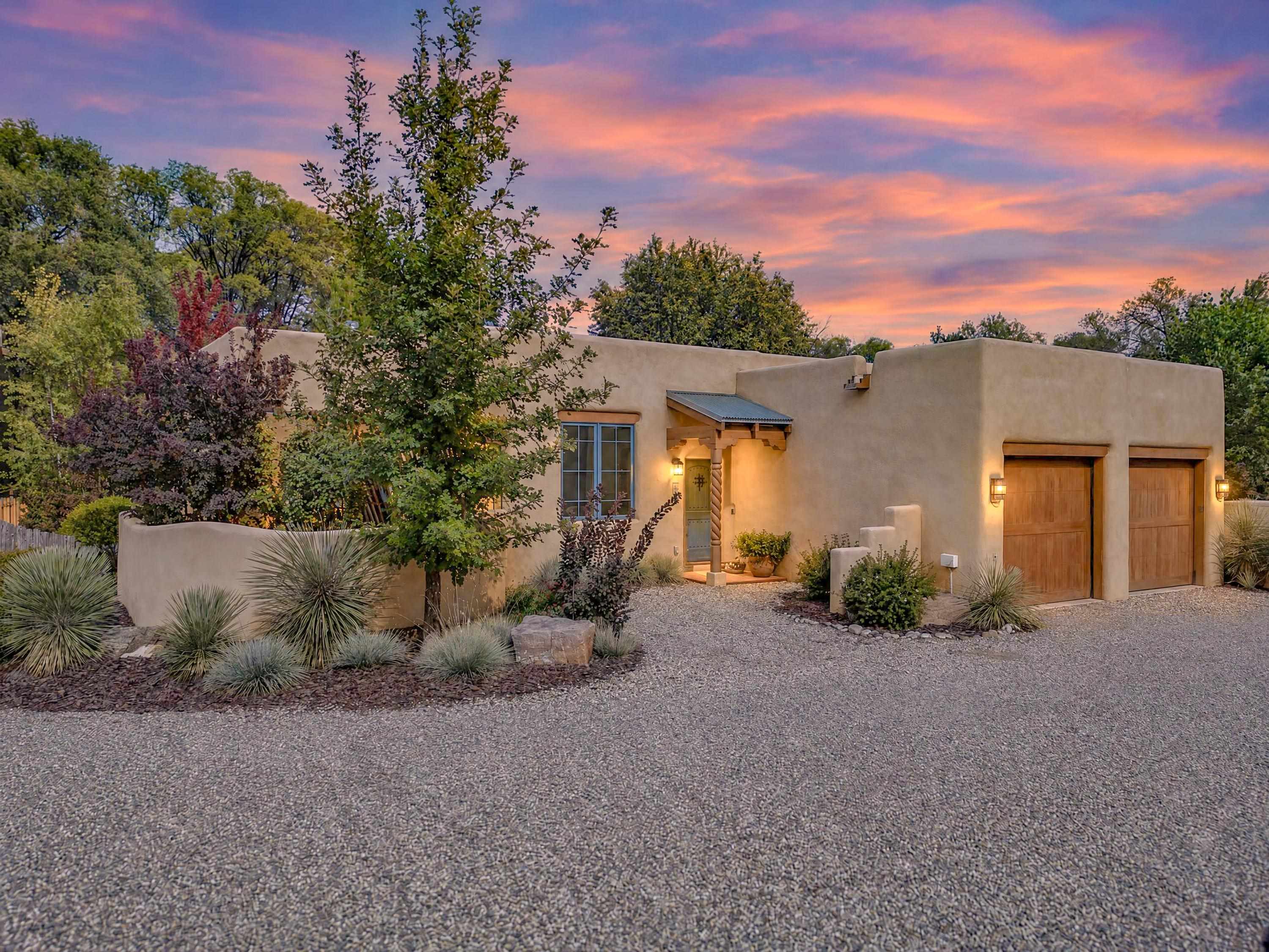 Adobe-style home with two wooden garage doors, gravel driveway, and desert landscaping at sunset.