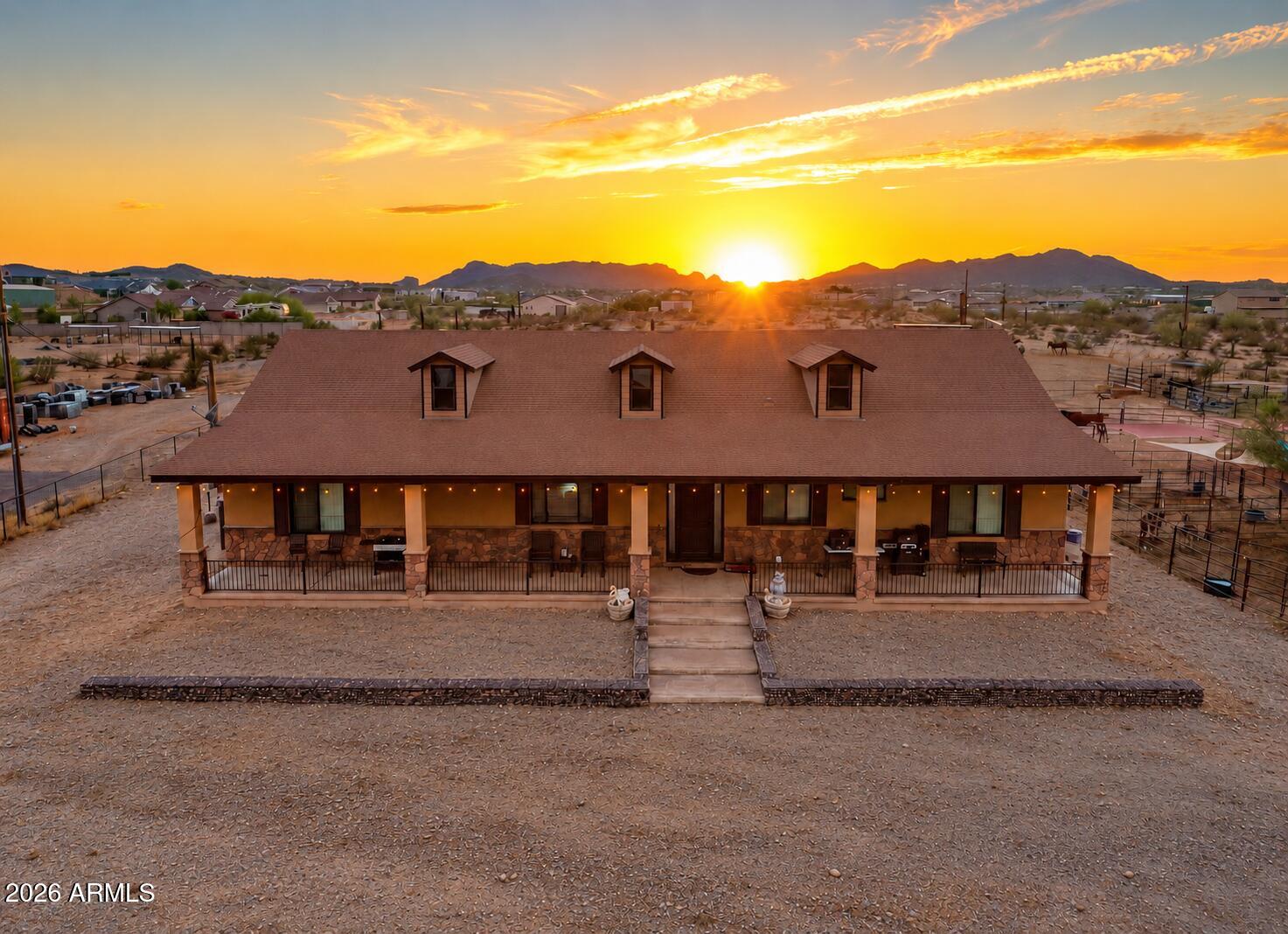 Desert home with a broad stone-fronted porch beneath a vivid sunset and mountain silhouettes.