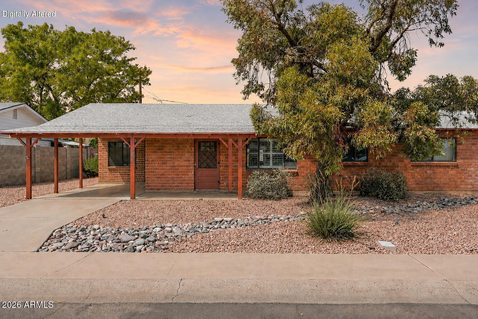 Red brick ranch house with a covered carport, desert rock landscaping, and mature trees at dusk.
