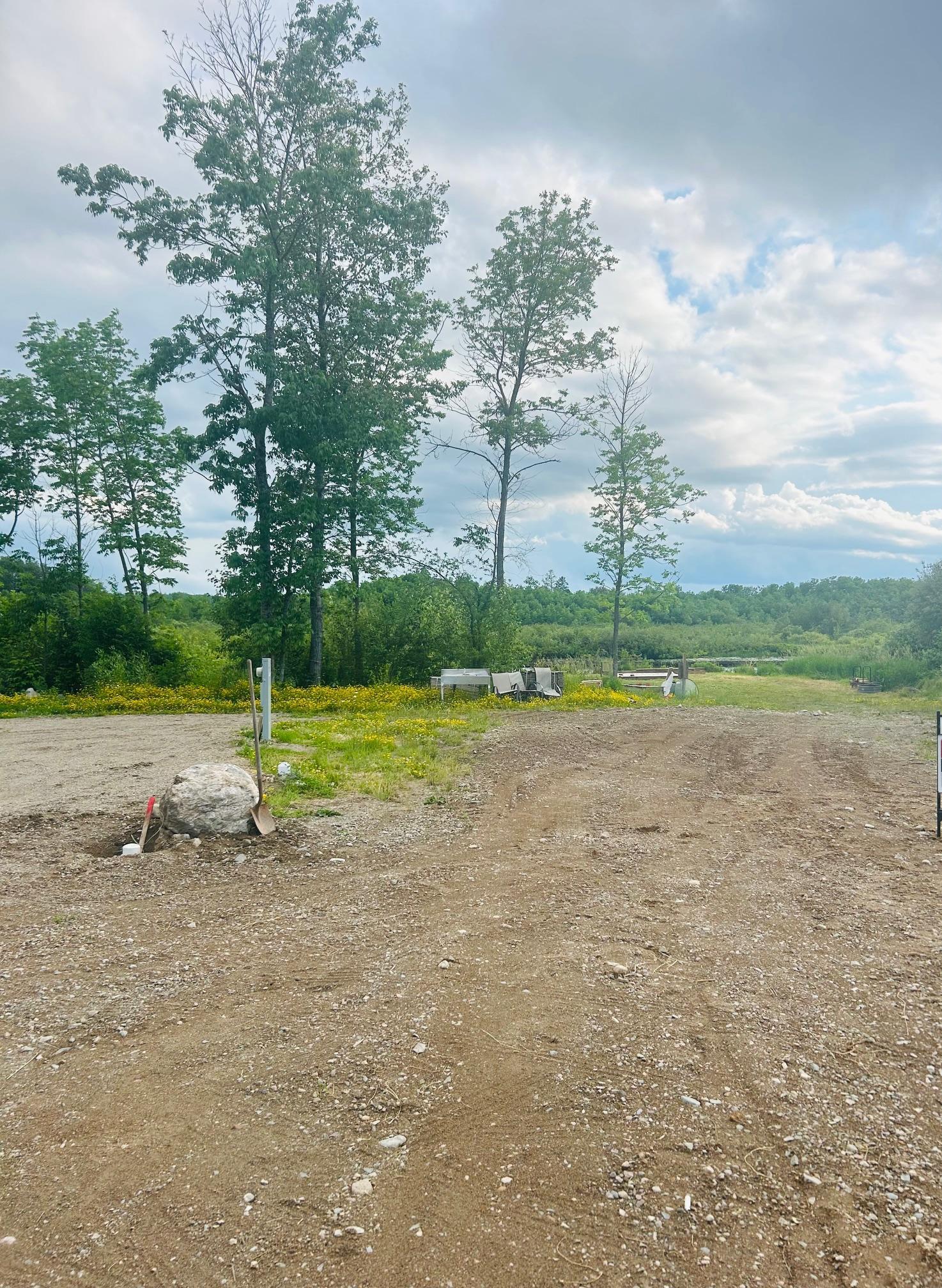 Cleared vacant lot with gravel ground, mature trees, outdoor chairs, and a glimpse of water.