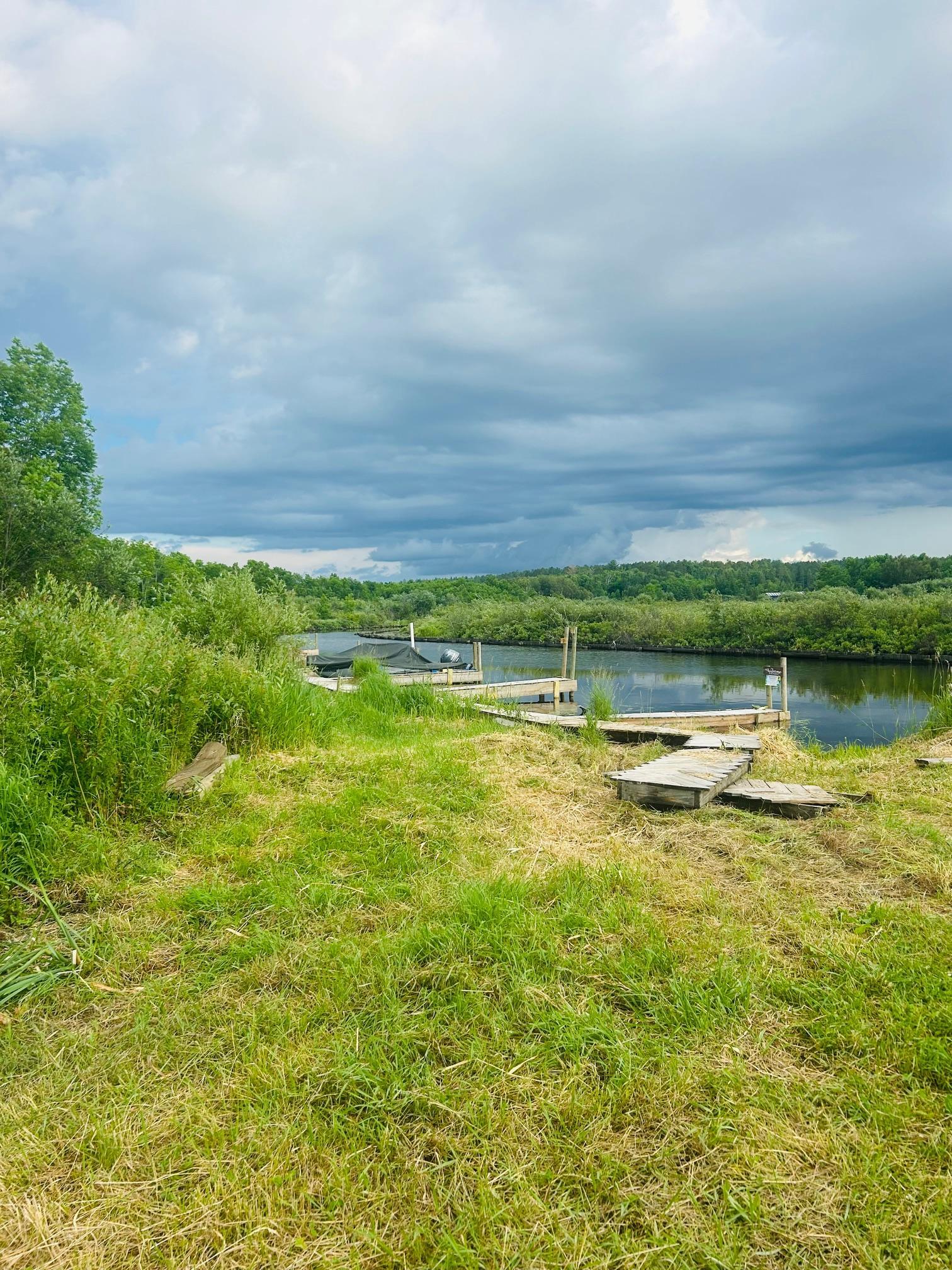 Waterfront lot with a wooden dock beside calm water and a tree-lined shore.