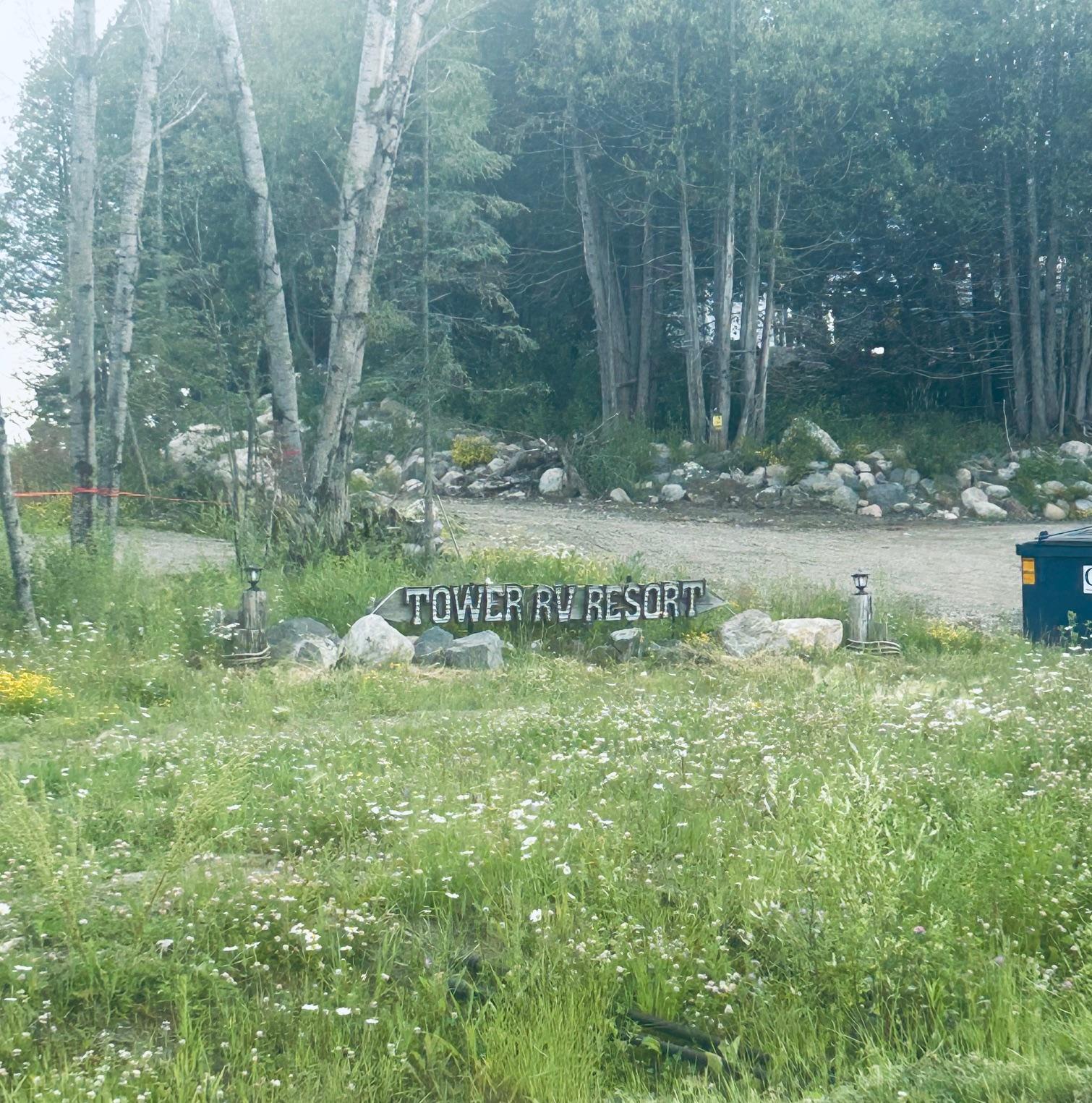 Wooded entrance to Tower RV Resort, with a gravel drive and a sign among rocks and wildflowers.