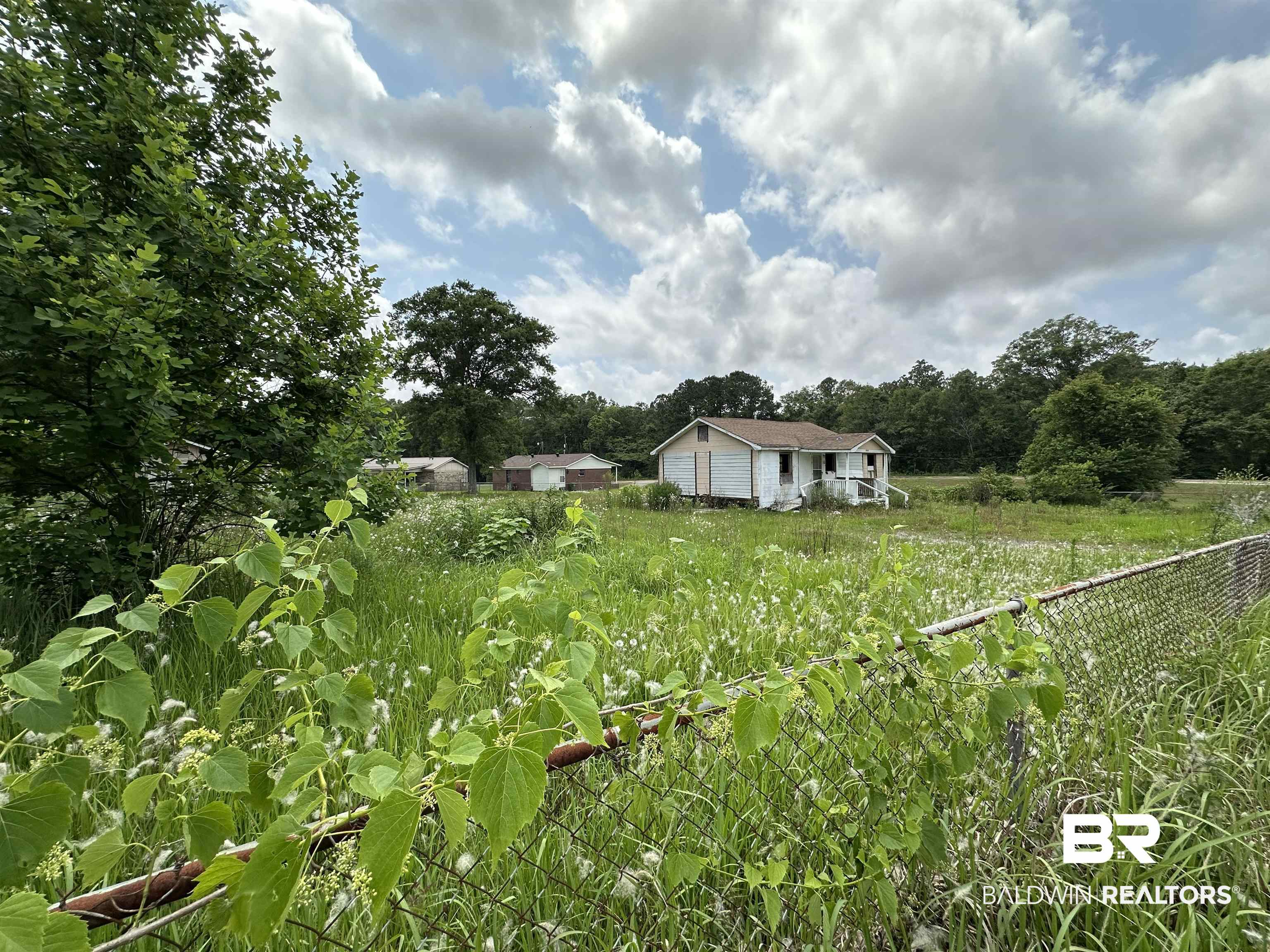 Small white house on an overgrown lot behind a chain-link fence.