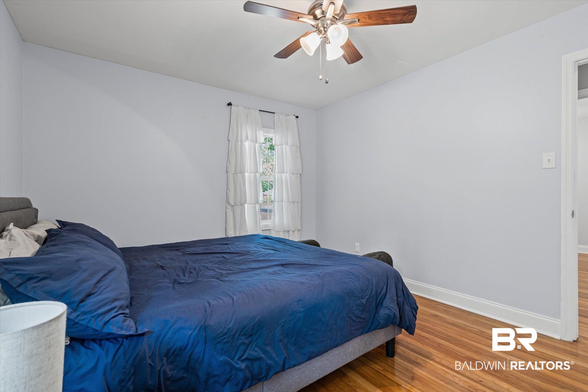 Bedroom with a navy-covered bed, hardwood floors, a curtained window, and a ceiling fan.