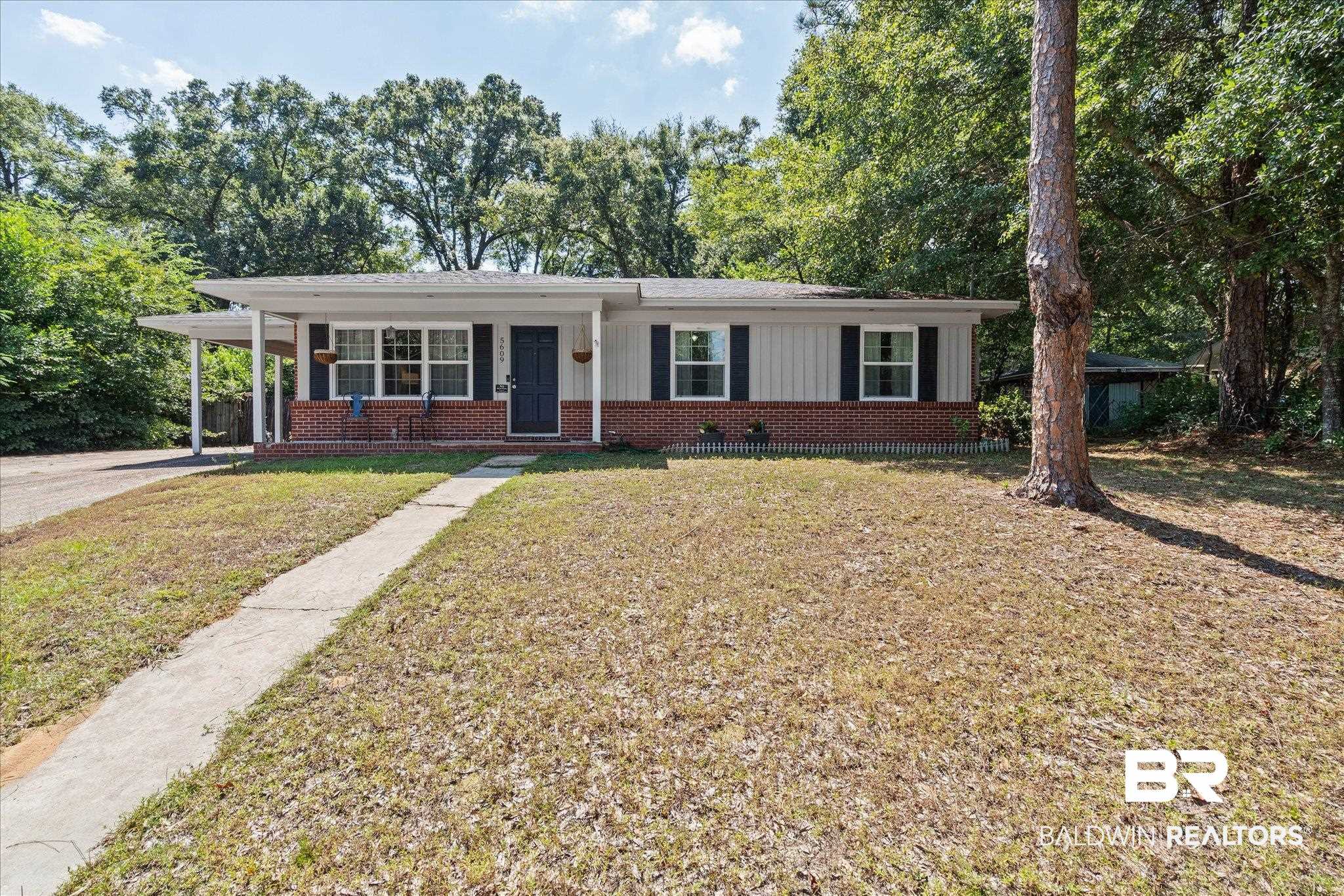 Single-story white-and-brick home with a covered carport, concrete walkway, and mature trees.