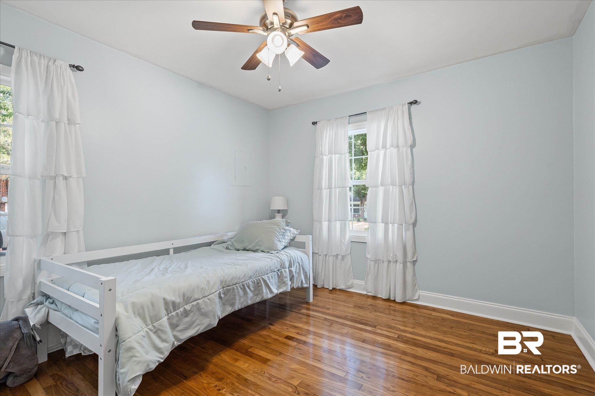 Bedroom with a white single bed, warm hardwood floors, and white-curtained windows.