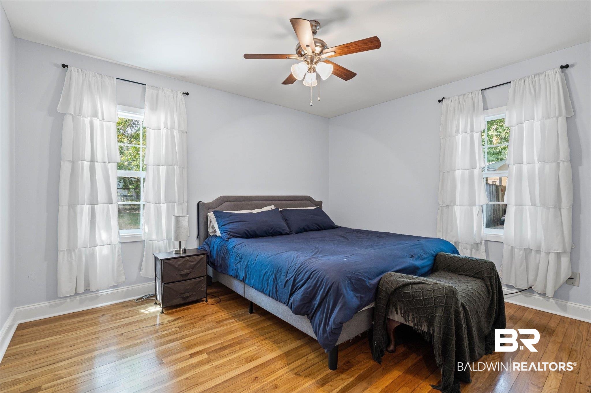 Bedroom with a blue bed, hardwood floors, two curtained windows, and a ceiling fan.