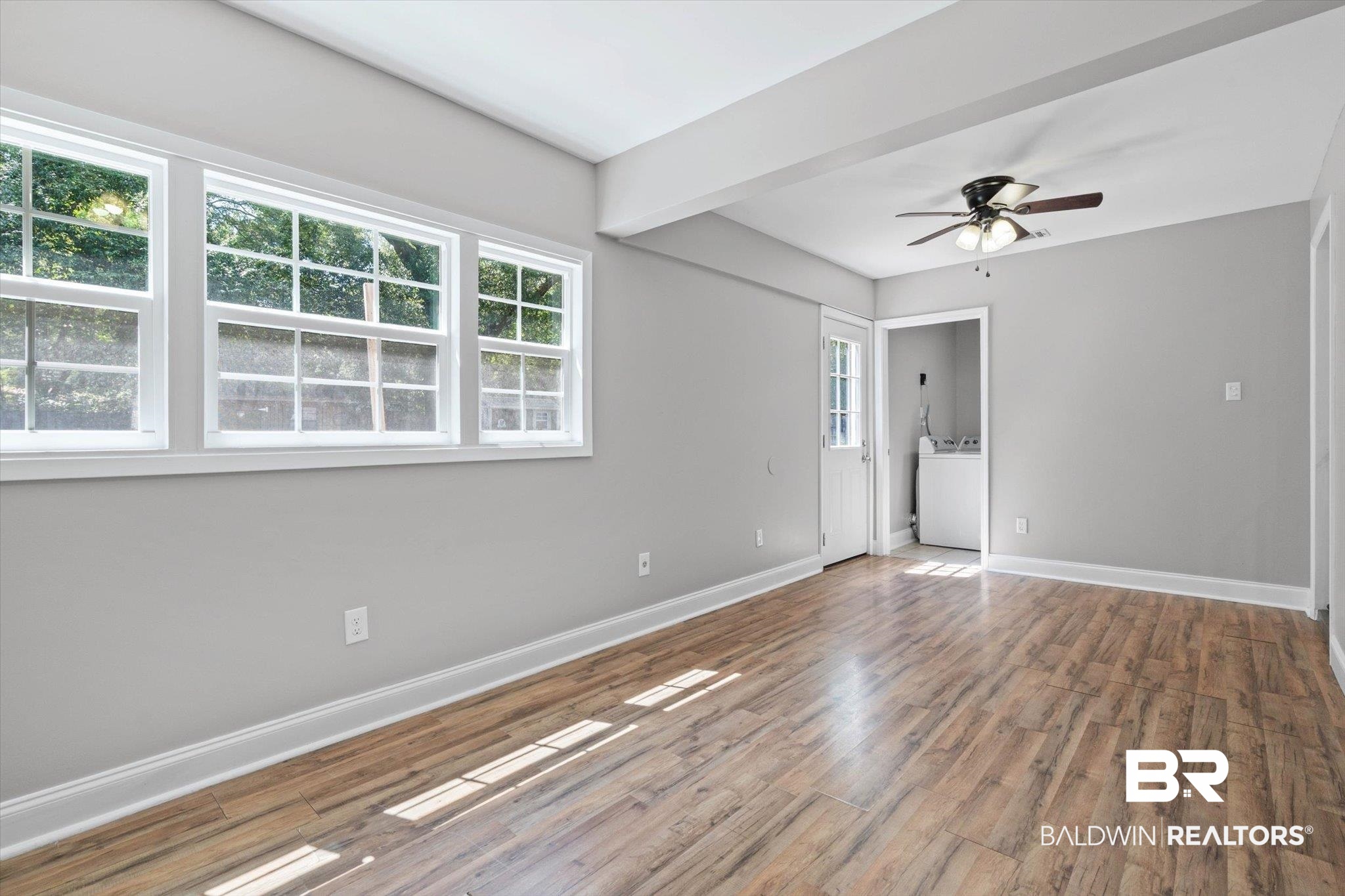 Vacant room with large windows, wood-look floors, a ceiling fan, and a washer visible through the doorway.