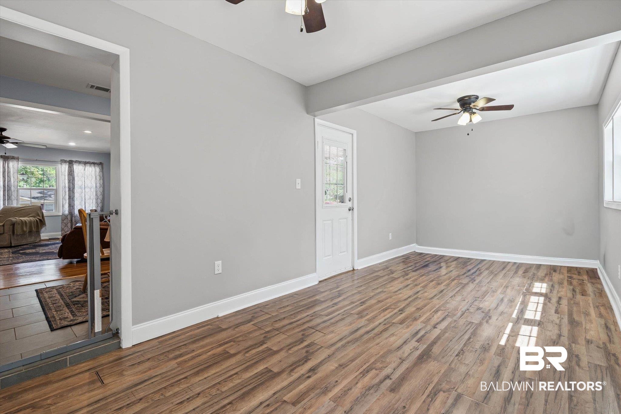 Open room with wood-look plank flooring, gray walls, ceiling fan, and a windowed door beside a furnished living area.