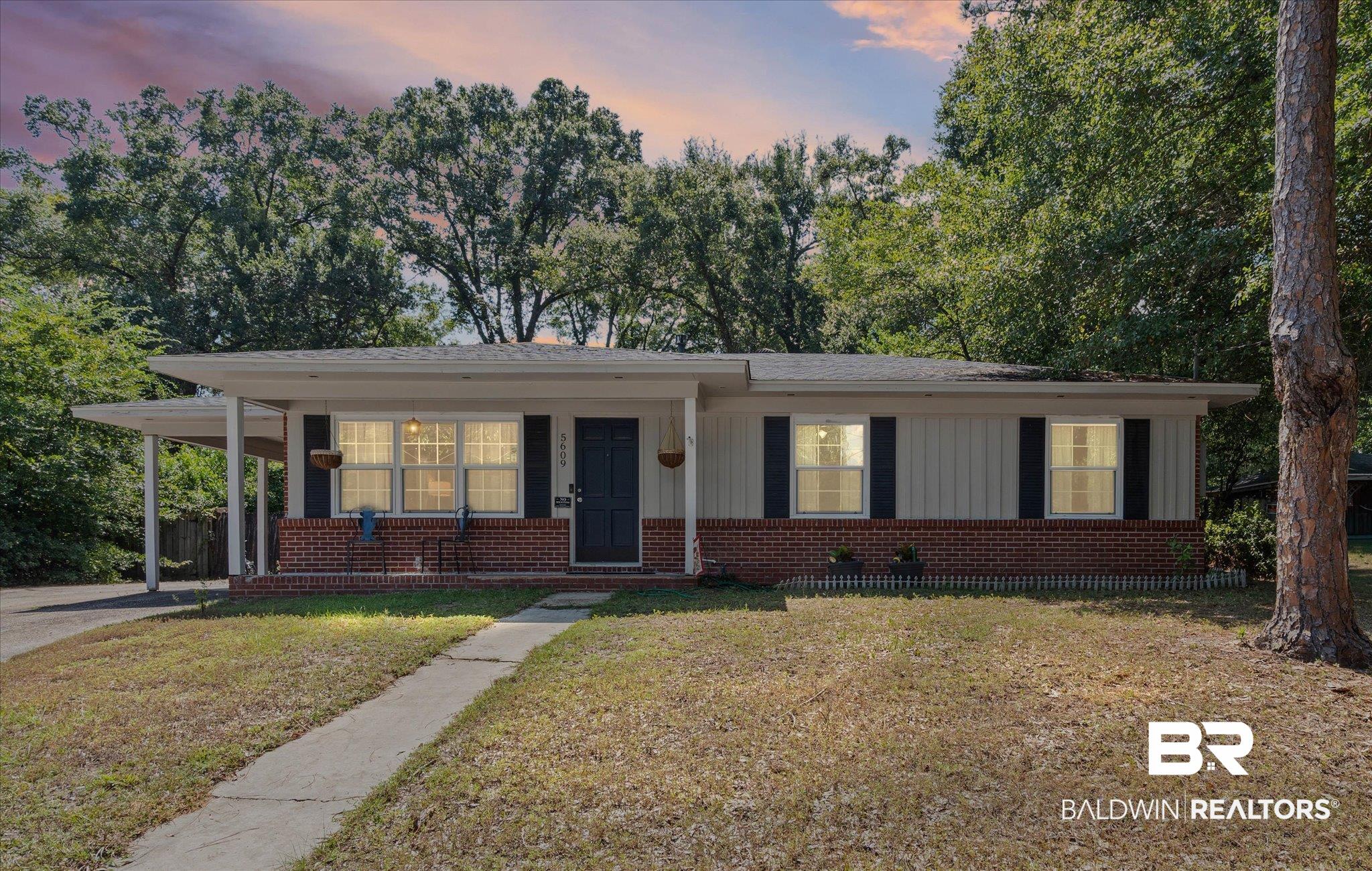 Single-story home with a red brick facade, covered porch, and carport beneath mature trees.