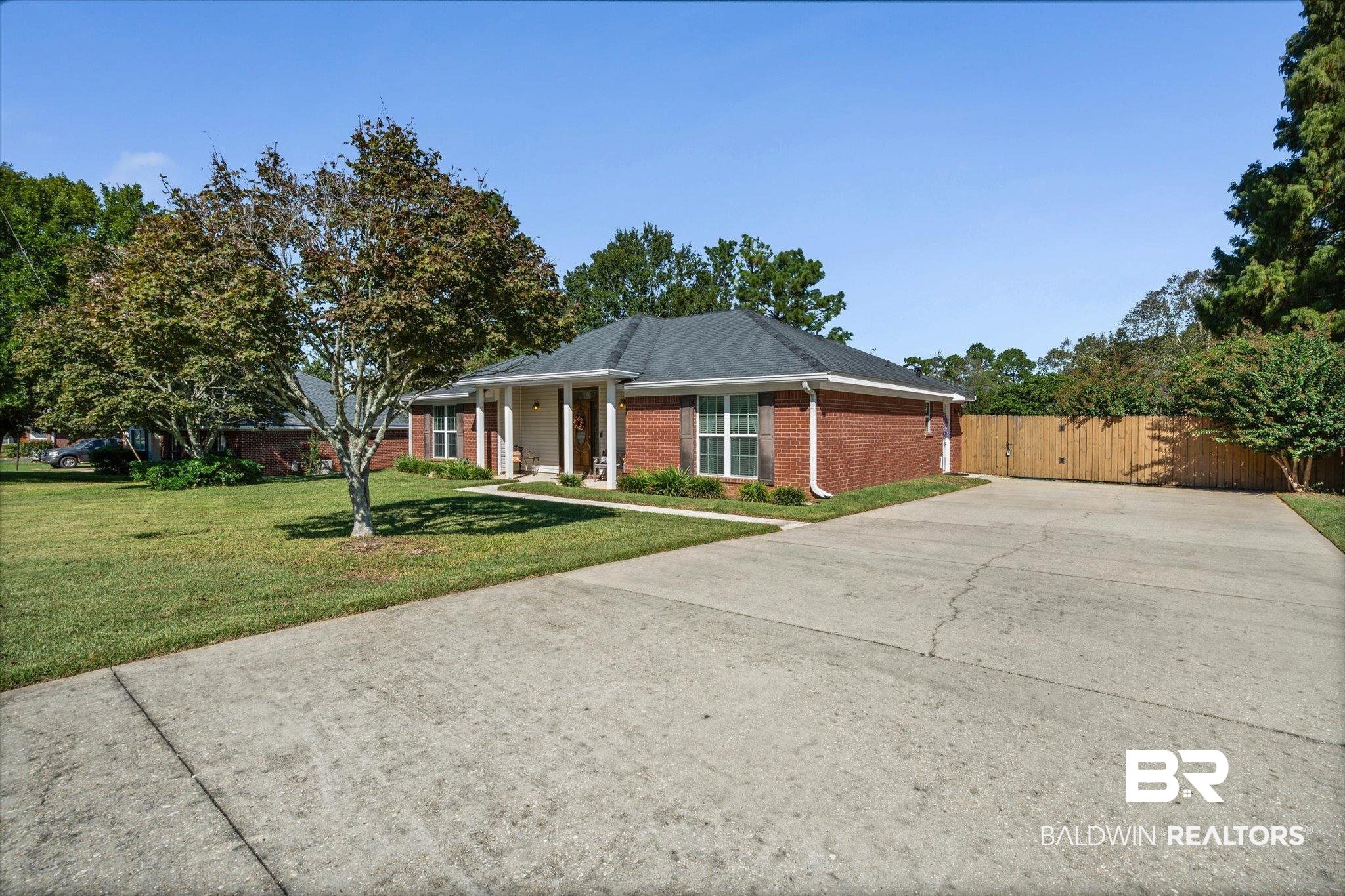 Red-brick single-story home with a covered entry, broad driveway, lawn, and wooden privacy fence.