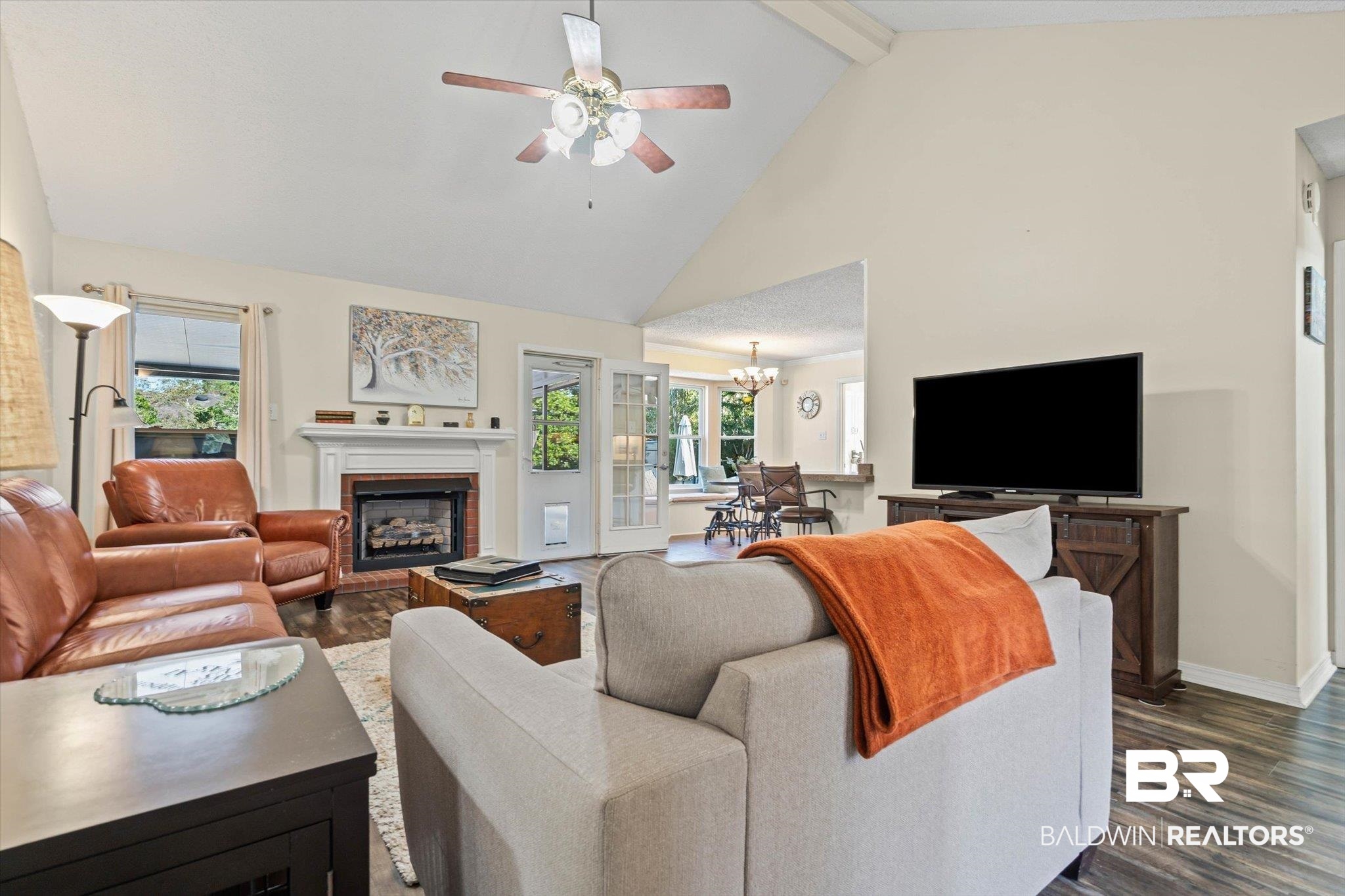 Living room with vaulted ceiling, brick fireplace, leather chairs, and a gray sofa with an orange throw.