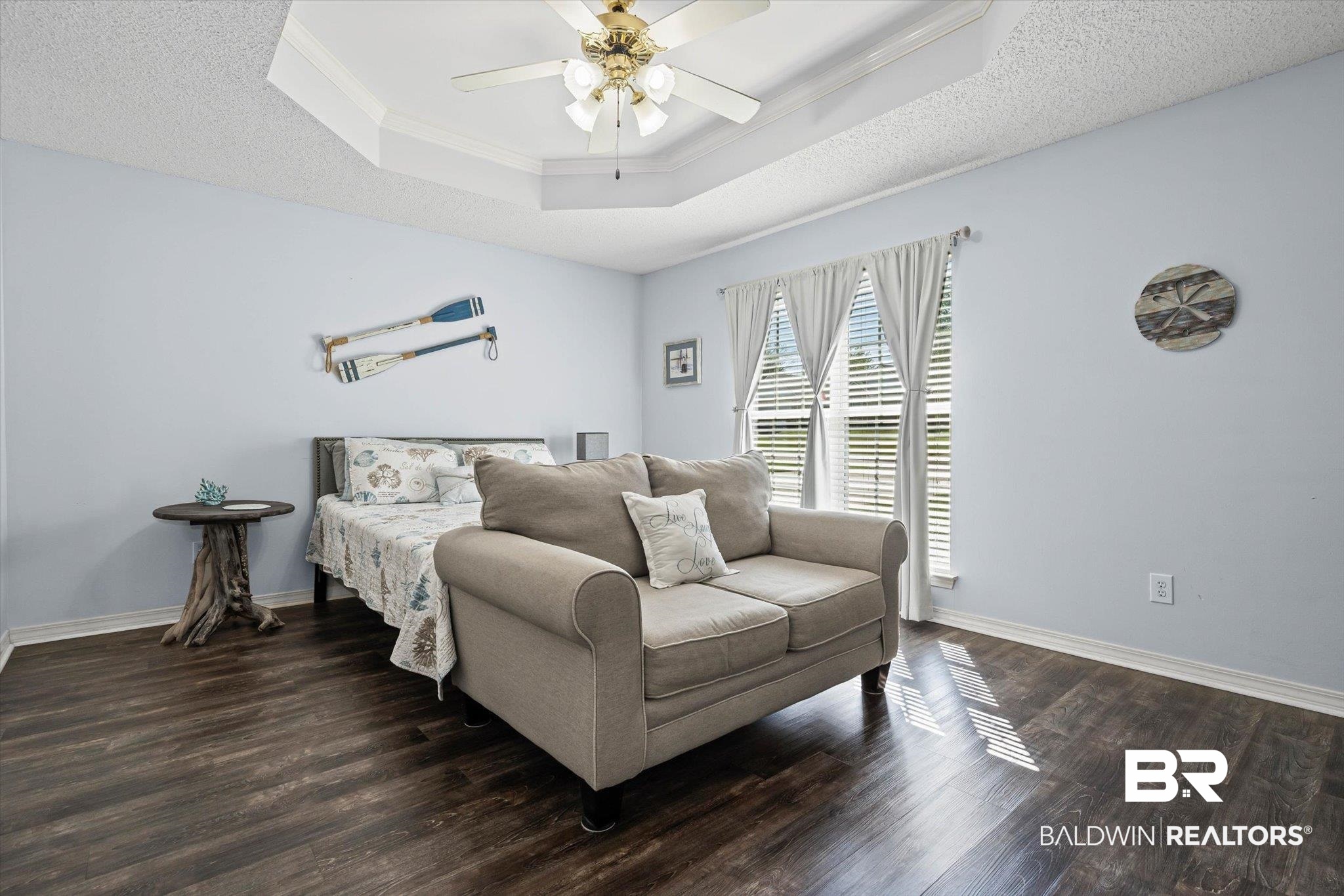 Bedroom with a beige loveseat in front of the bed and sunlight across dark wood-look flooring.