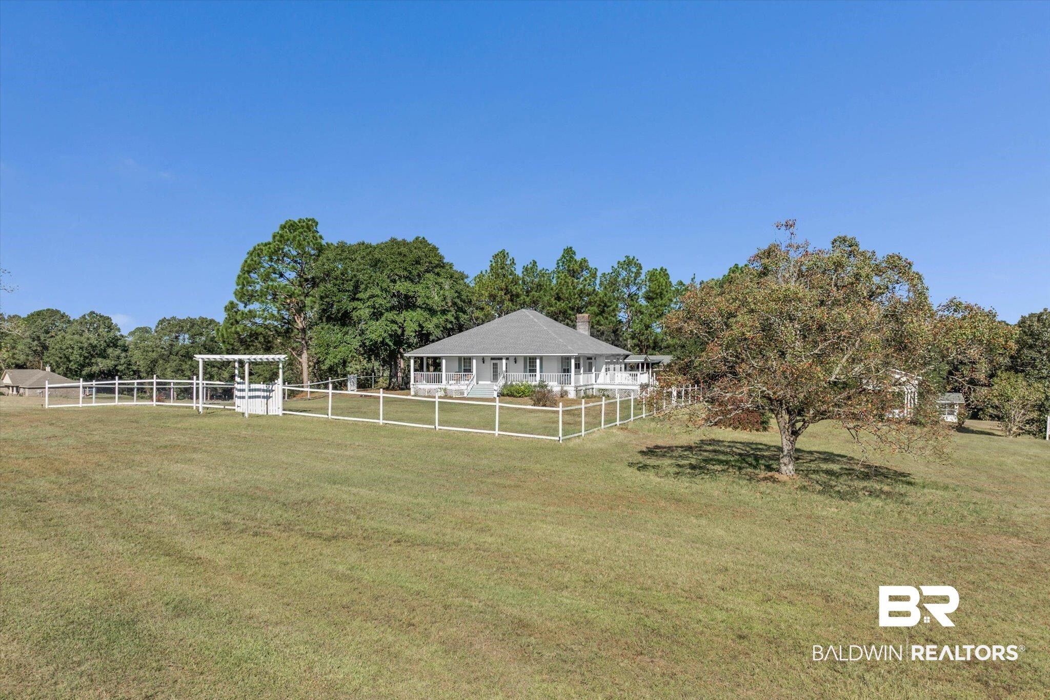 White single-story home with wraparound porch, fenced lawn, and pergola on a spacious tree-lined lot.