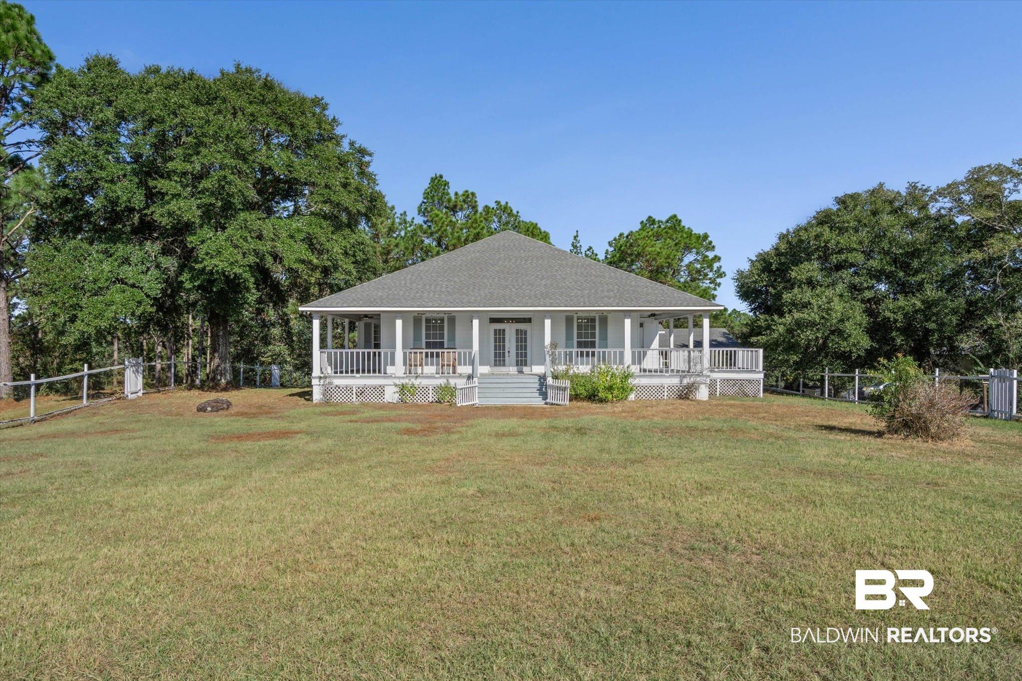 Single-story home with a broad covered porch, centered in a fenced grassy yard surrounded by mature trees.