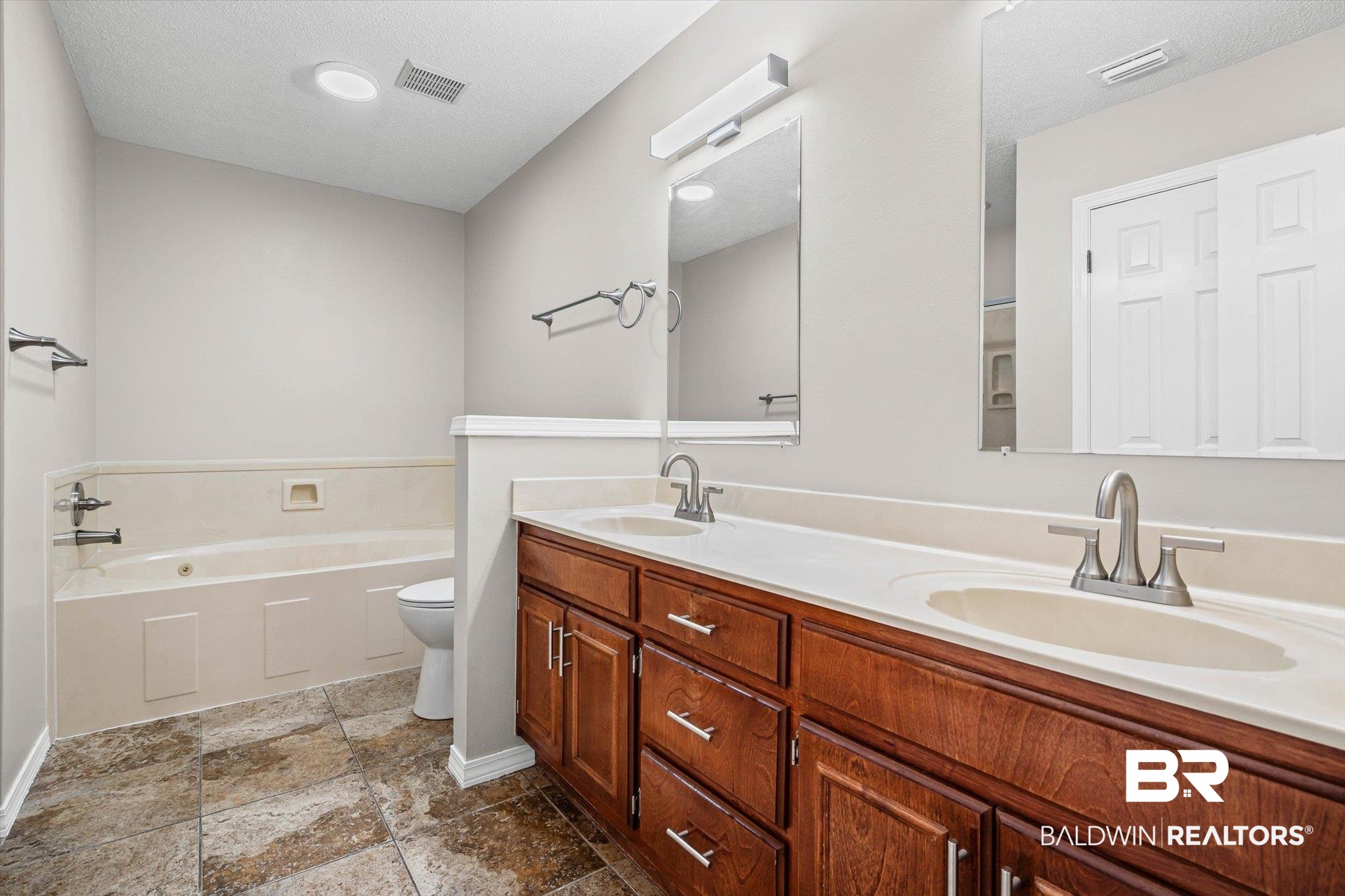Bathroom with a double-sink wood vanity, bathtub, and stone-patterned tile floor.