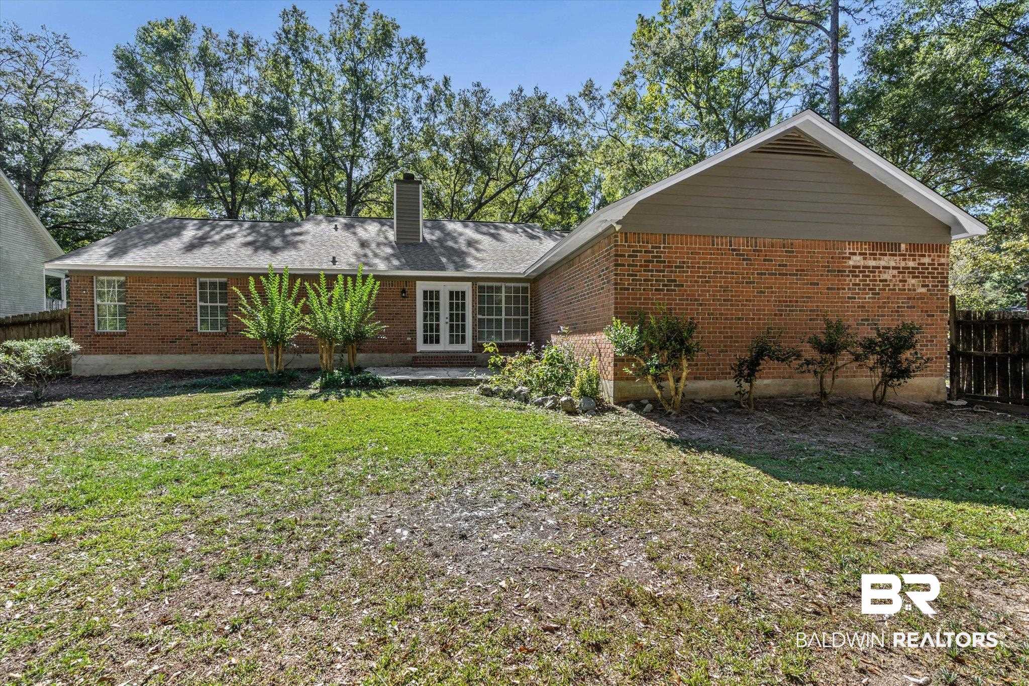 Red-brick home with white French doors and a broad, patchy backyard shaded by mature trees.