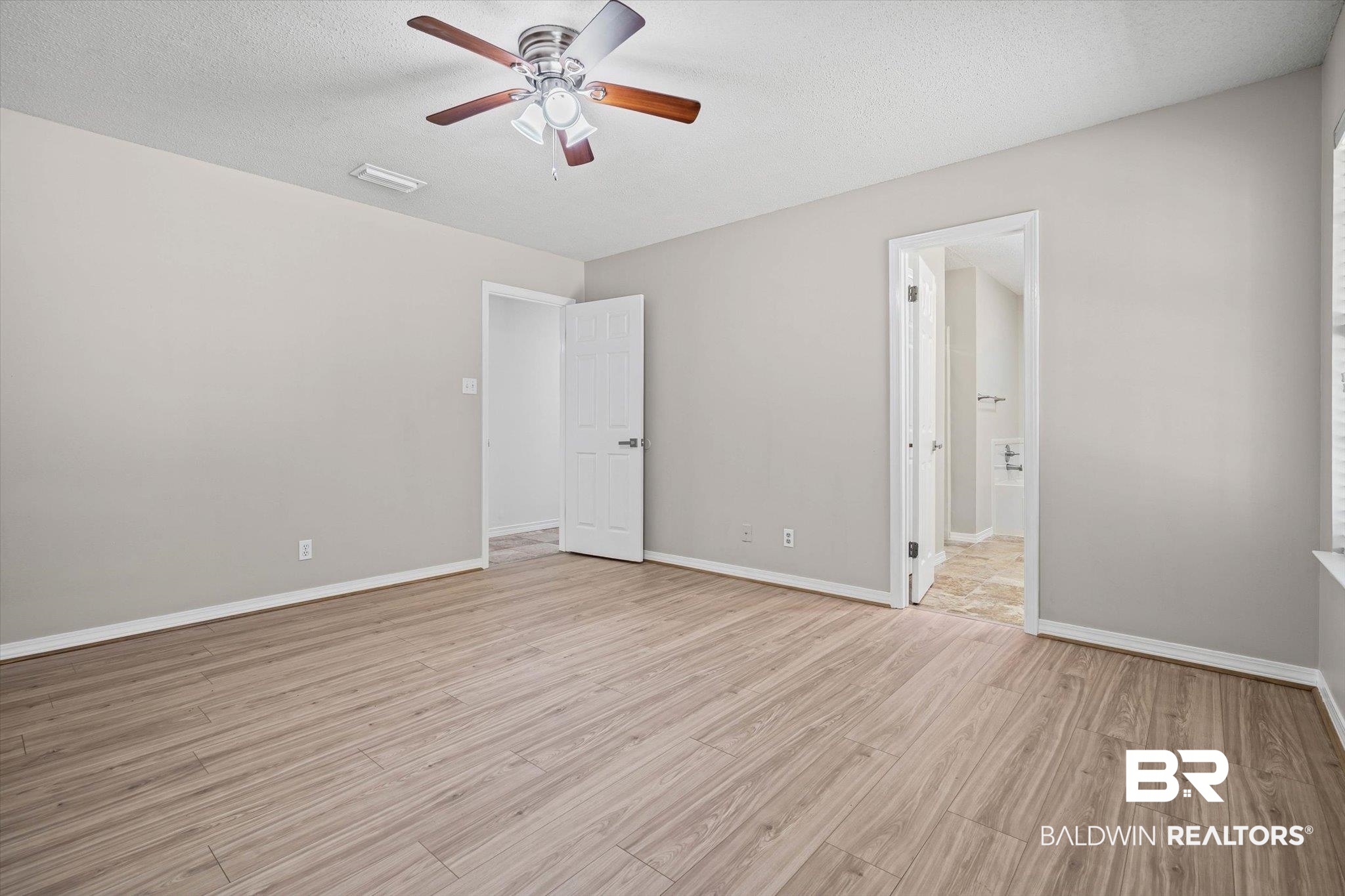 Vacant bedroom with wood-look flooring, a ceiling fan, and an open doorway to a tiled bathroom.