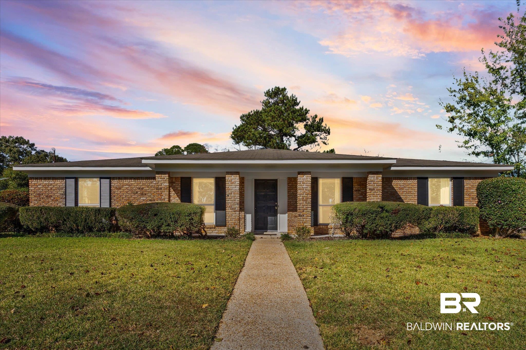 Single-story brick house with trimmed hedges and a straight front walkway beneath a colorful sunset sky.