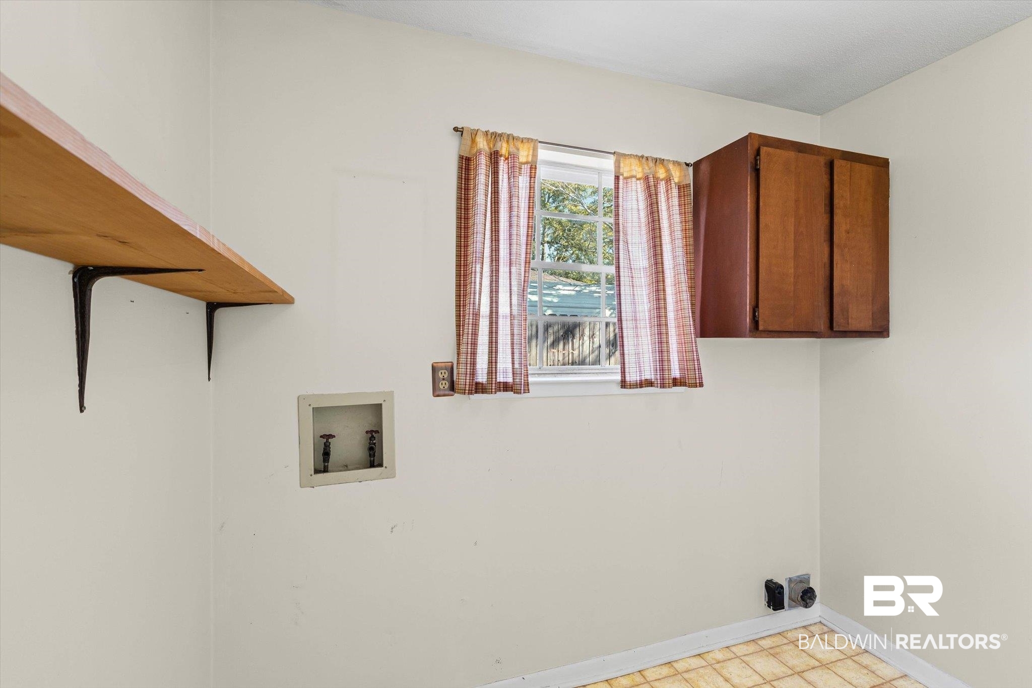 Empty laundry room with washer and dryer hookups, a wooden shelf, wall cabinet, and curtained window.