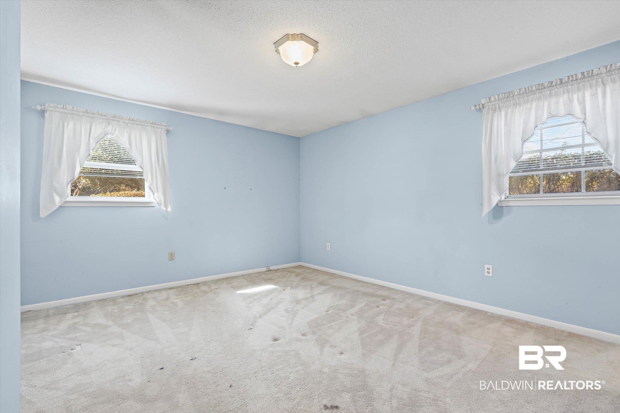 Empty light-blue room with beige carpet, two curtained windows, and a ceiling light.
