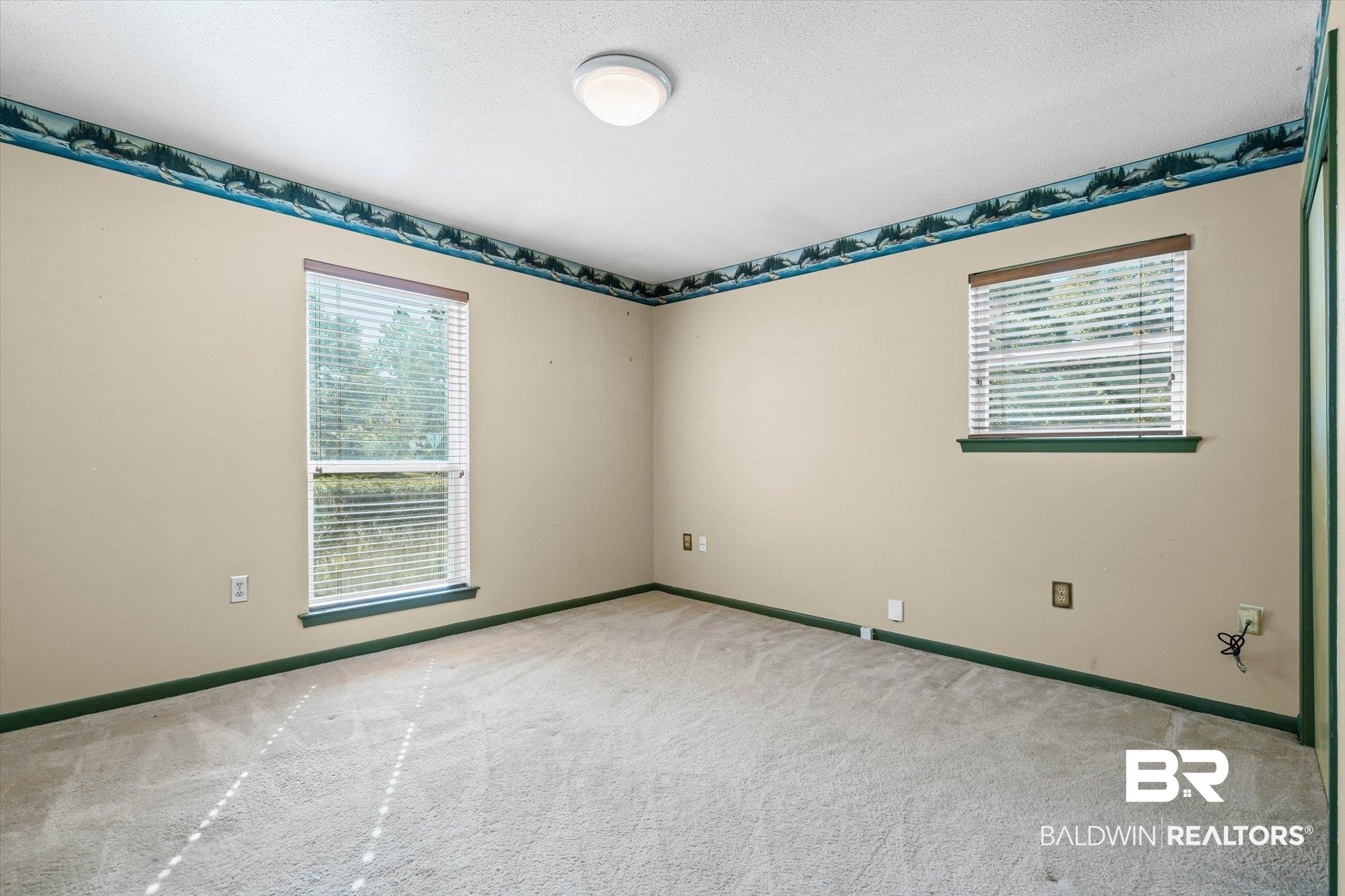 Empty carpeted room with beige walls, green trim, and two windows with blinds.