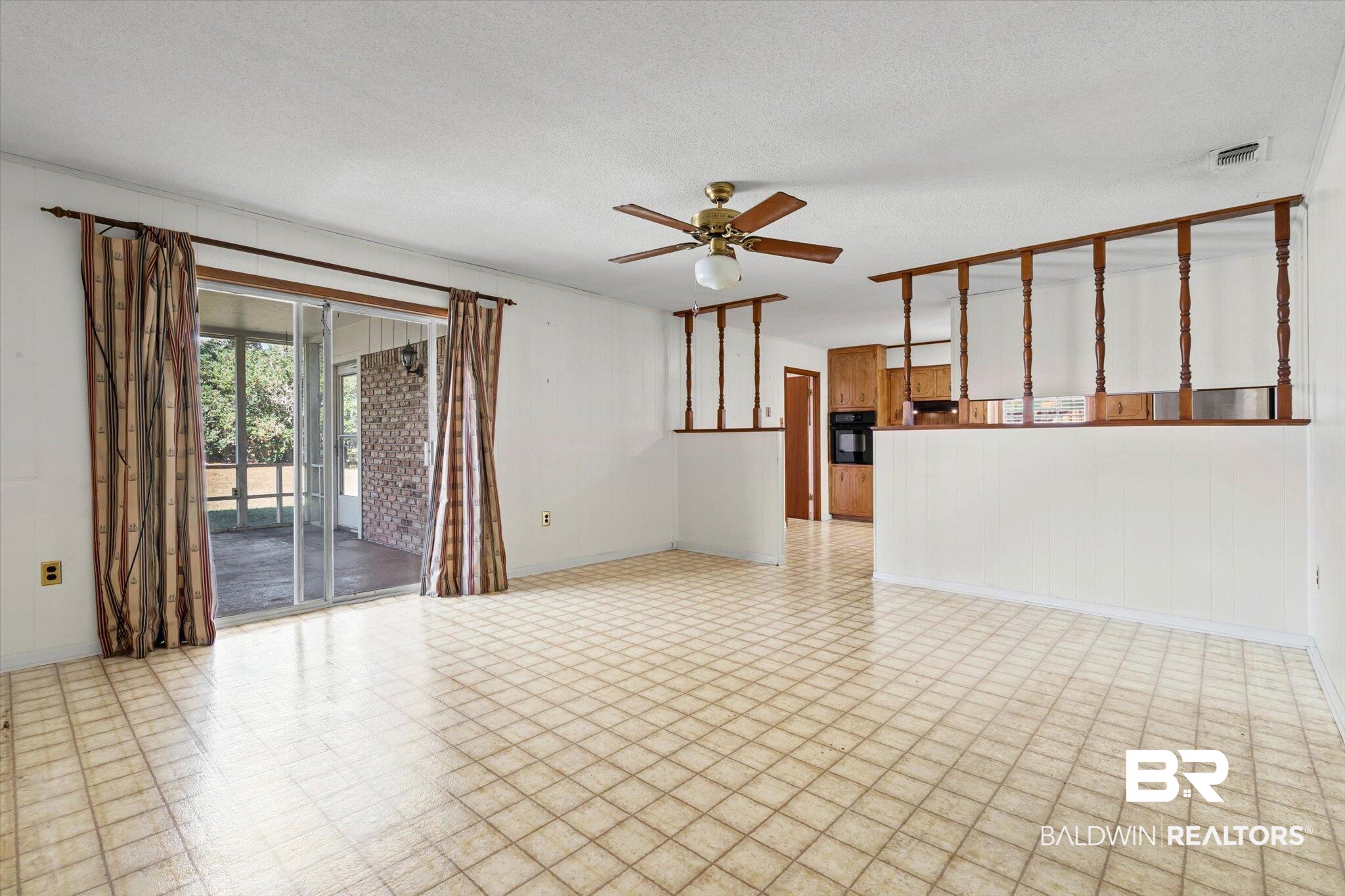 Empty tiled living area with ceiling fan, sliding doors to a porch, and a raised kitchen pass-through.