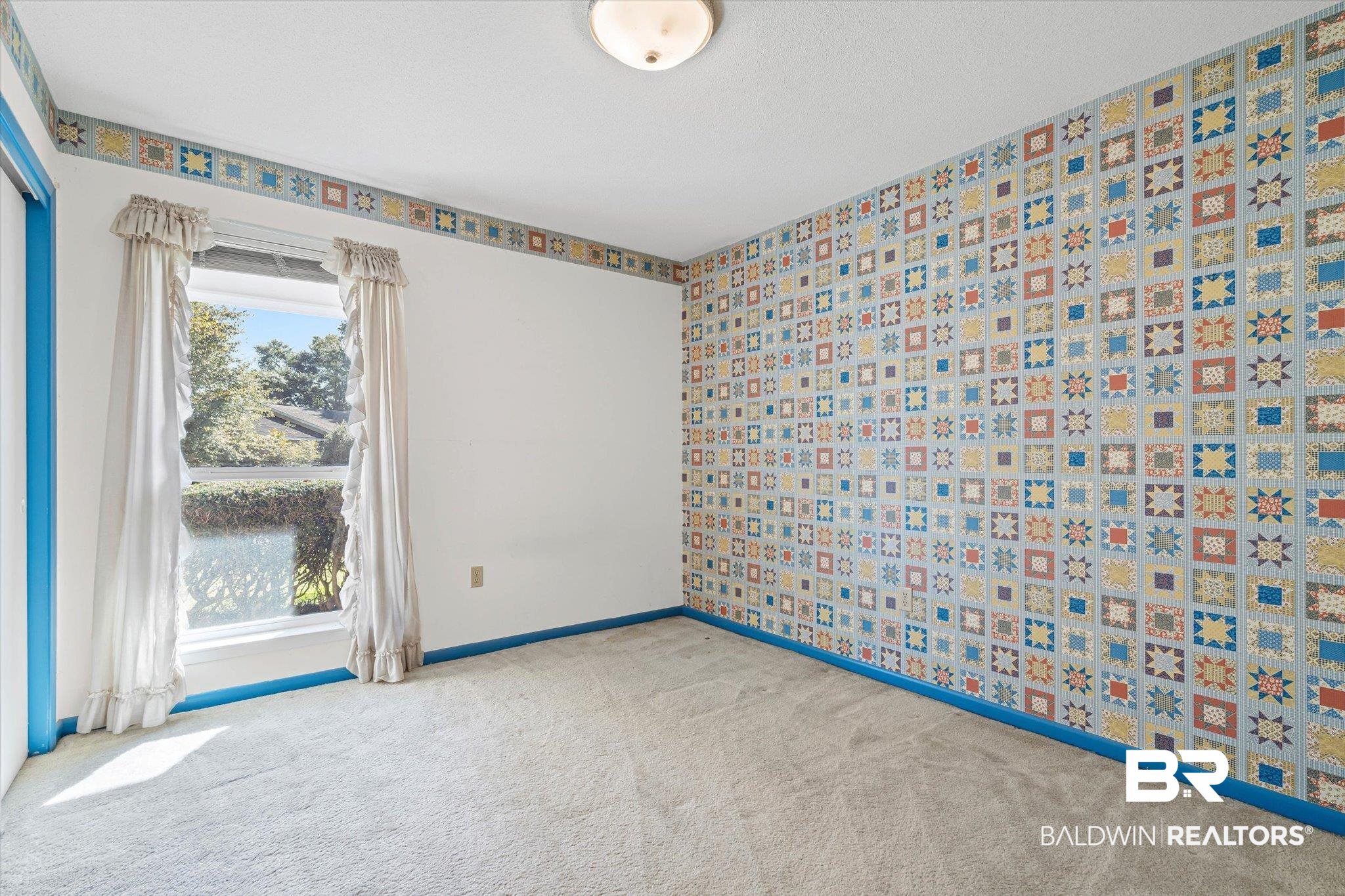 Empty bedroom with colorful patchwork wallpaper, bright blue trim, and a curtained window.