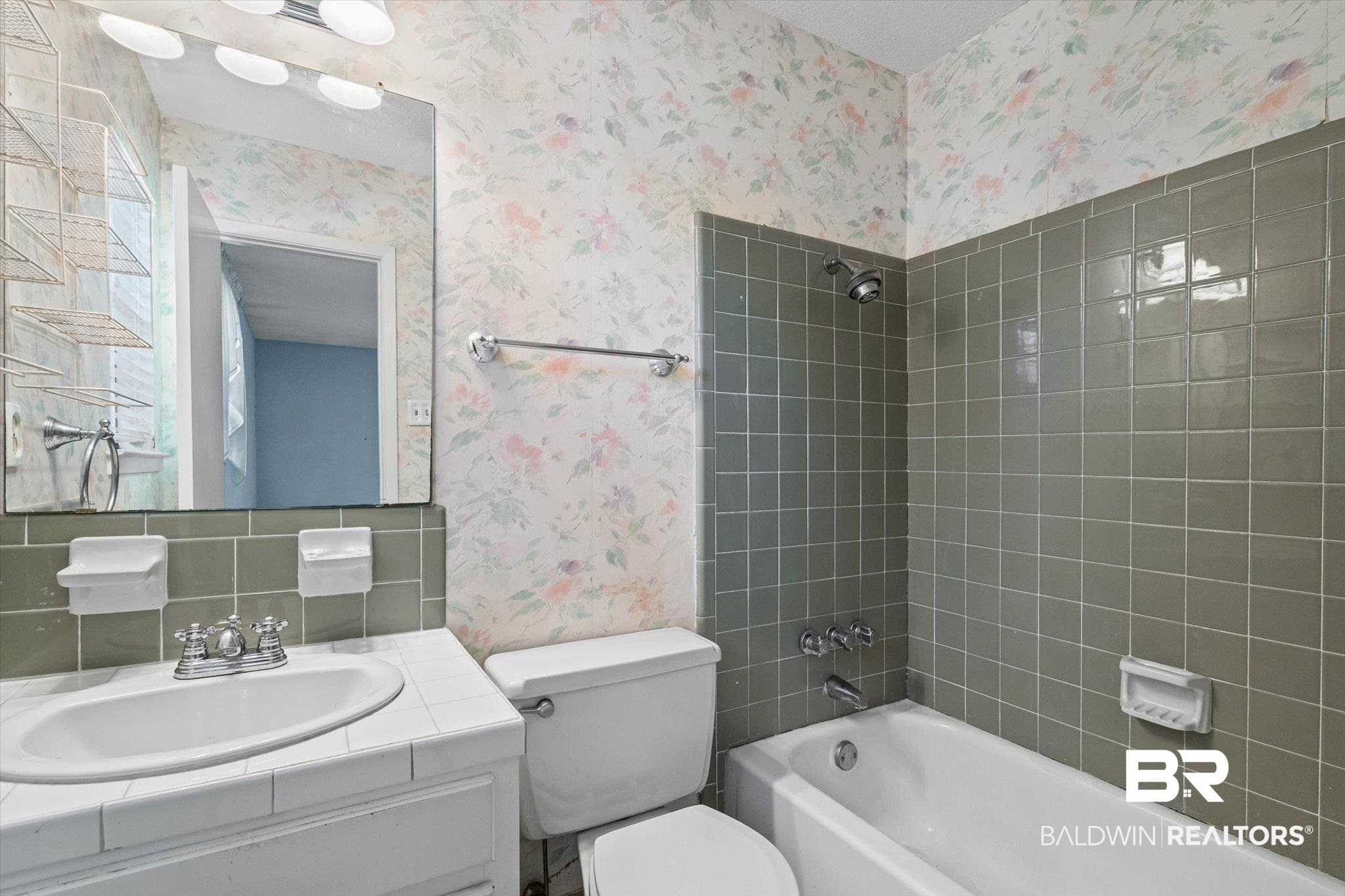 Bathroom with gray-green tiled tub surround, floral wallpaper, white toilet, and tiled vanity.
